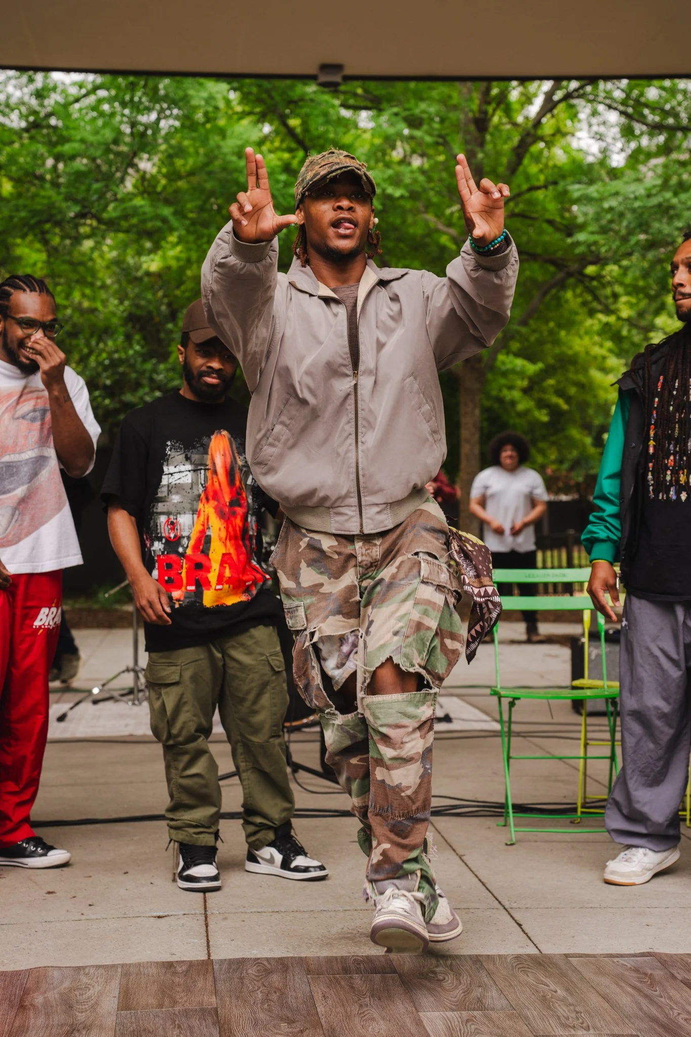 A group of people standing outdoors with trees in the background. The central person, wearing a beige jacket, camouflage pants, and a cap, is dancing or performing. Others are watching, some with smiles, in casual clothing.