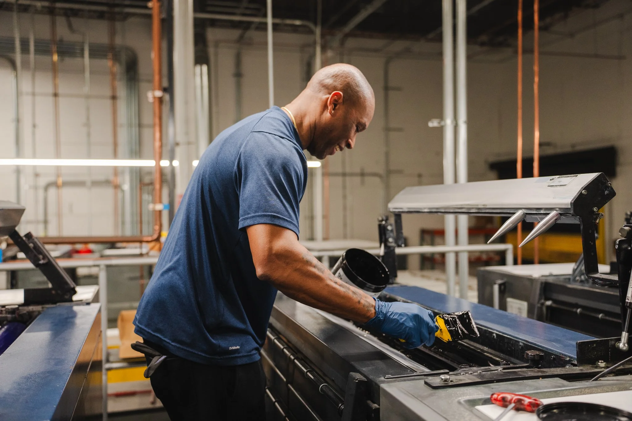 A man working in an industrial setting, wearing gloves and a blue shirt, inspecting or assembling a component at a metal workstation with machinery and pipes in the background.