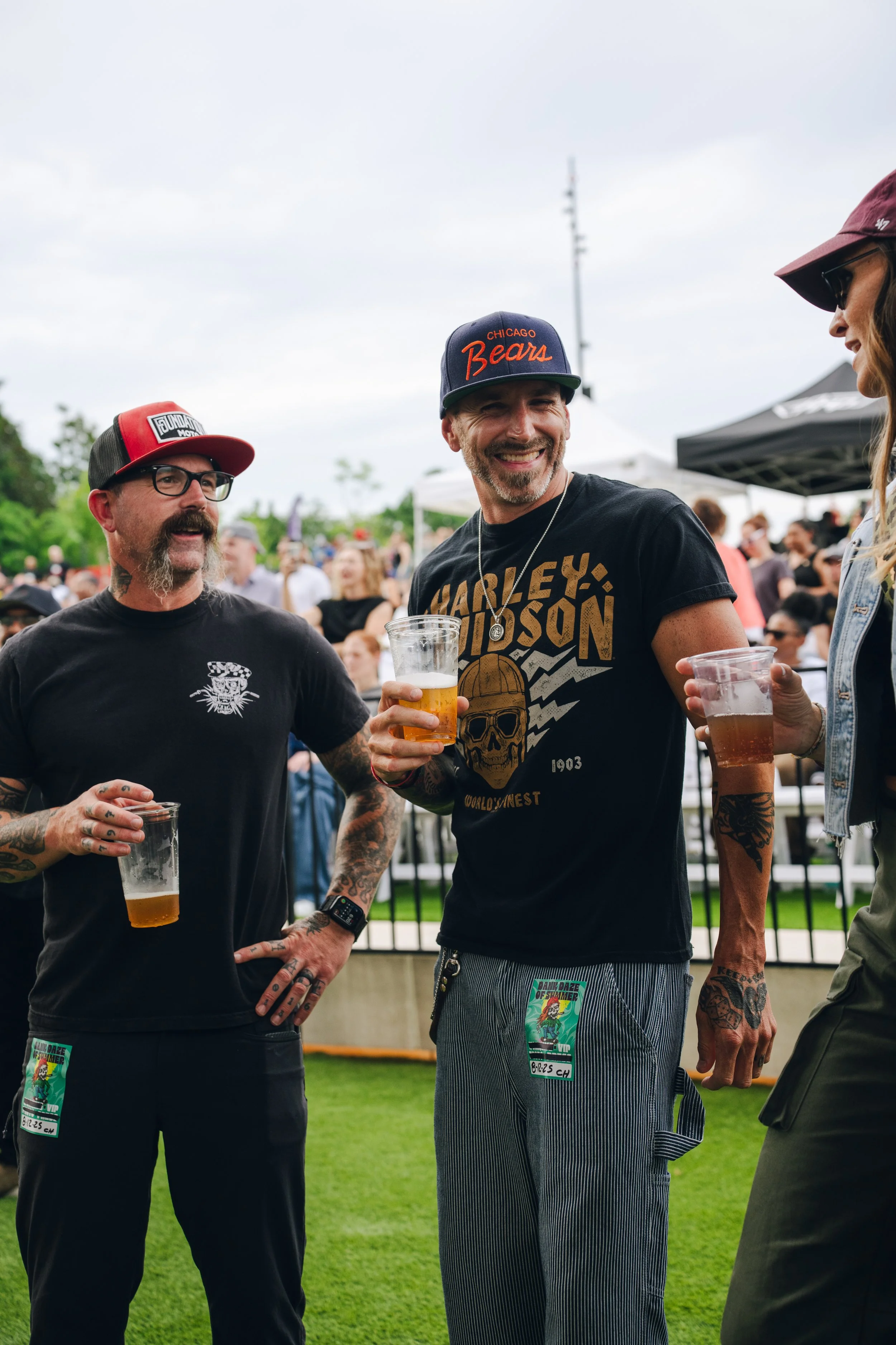 Three people are enjoying a gathering outdoors, holding drinks and chatting. The man in the middle is smiling, wearing a black t-shirt with a skull graphic, a Chicago Bears cap, and has tattoos. The man on the left has a beard, glasses, a red and bla
