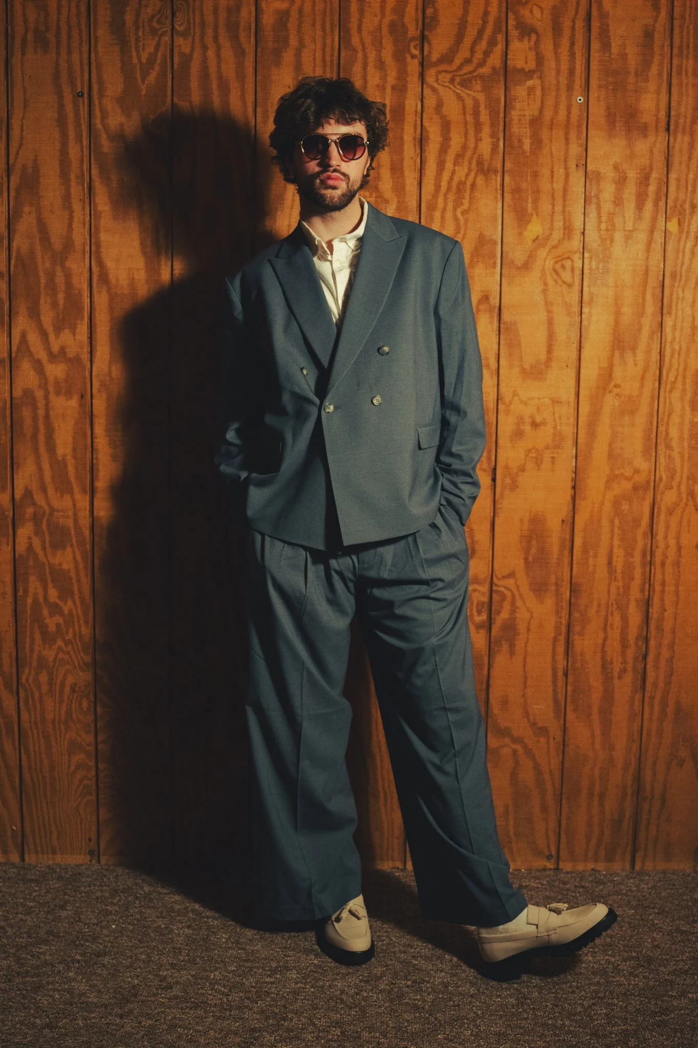 A man in a gray suit with sunglasses stands against a wooden wall with one hand in his pocket. He is wearing a white shirt, beige boots, and beige shoes. The setting has warm lighting and a textured carpet on the floor.