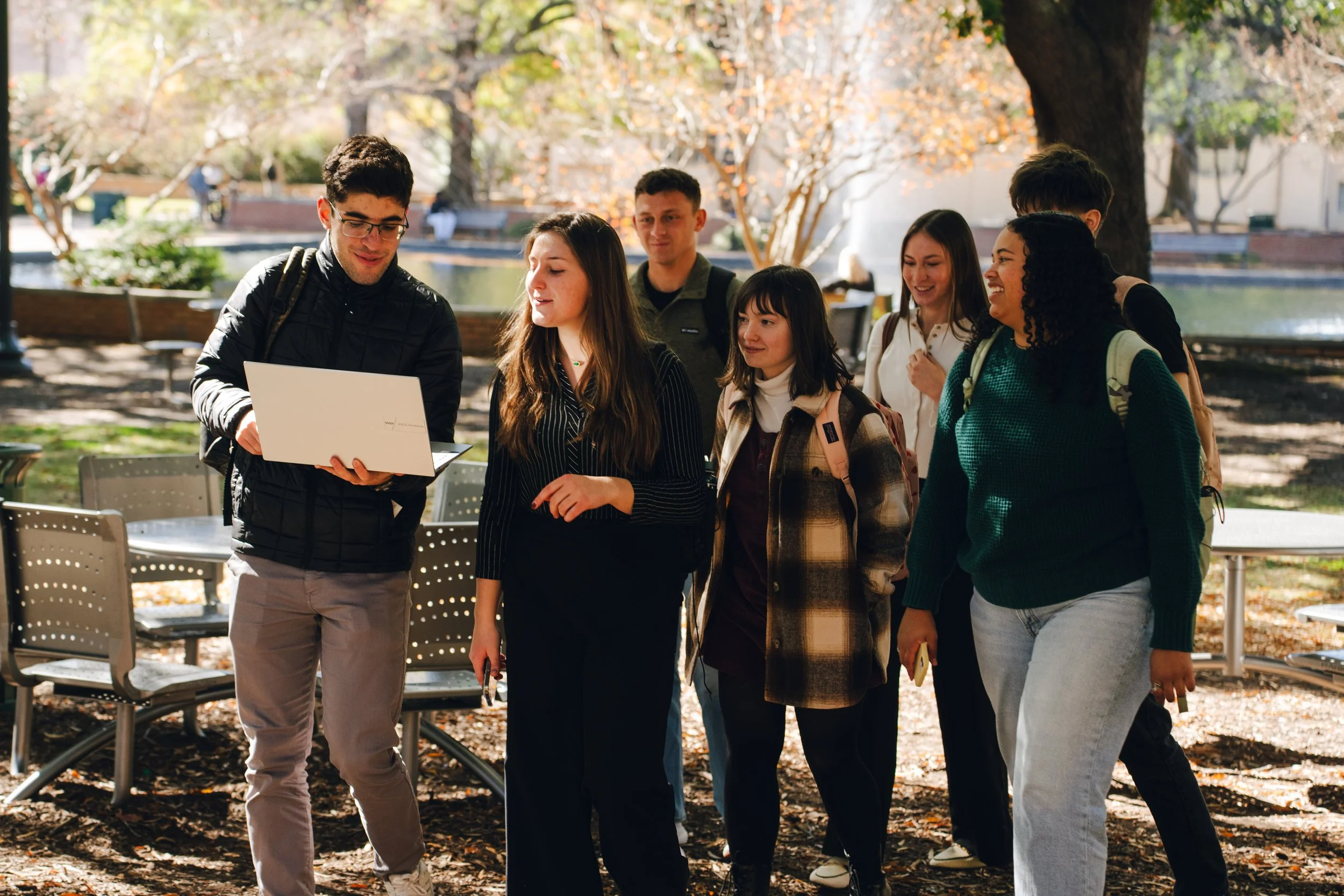 A group of young adults standing outdoors in a park, looking at a laptop held by a person wearing glasses and a black jacket, during autumn with trees and benches visible in the background.