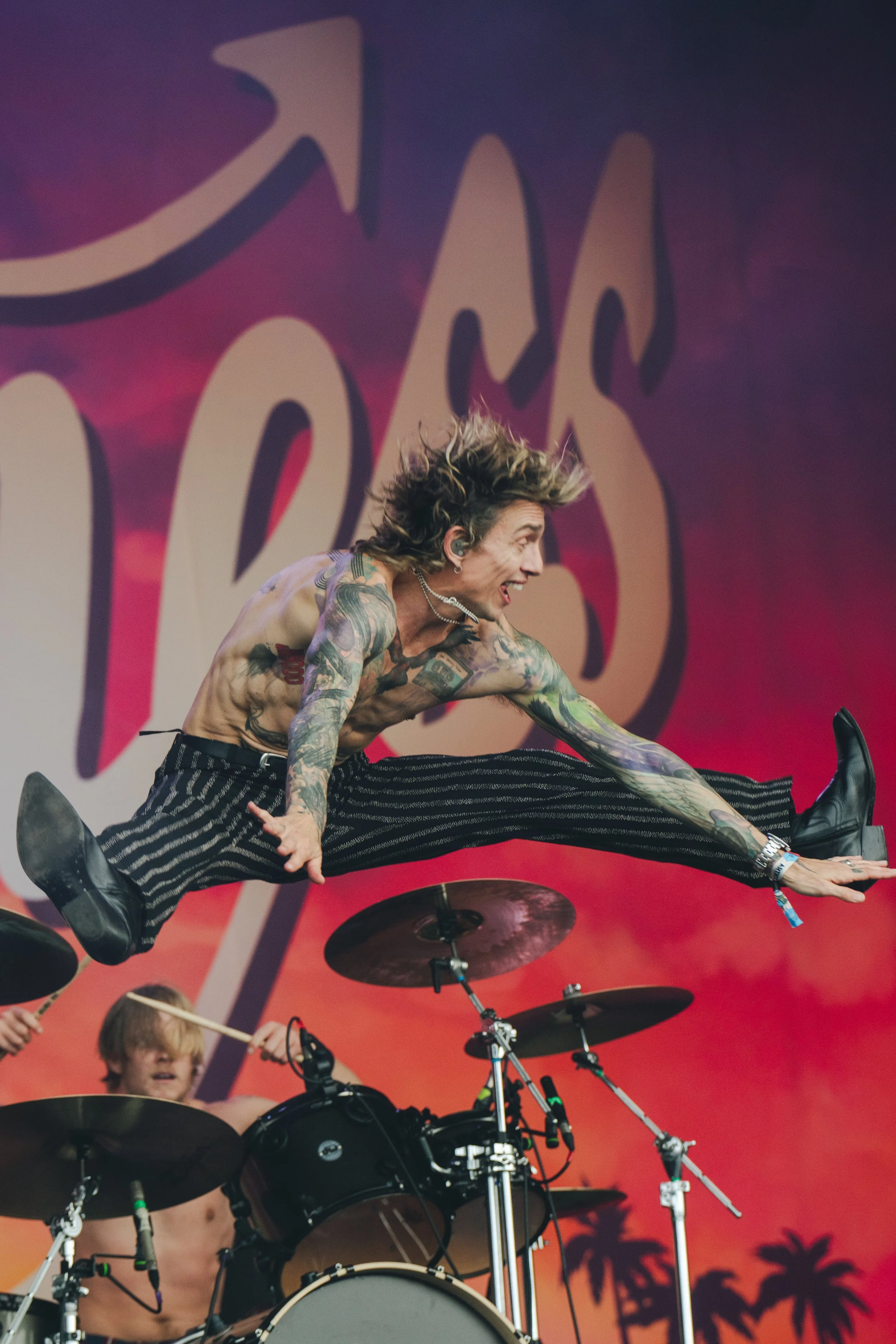 A shirtless male drummer performing on stage with a drummer in the background against a sunset backdrop with silhouettes of palm trees.