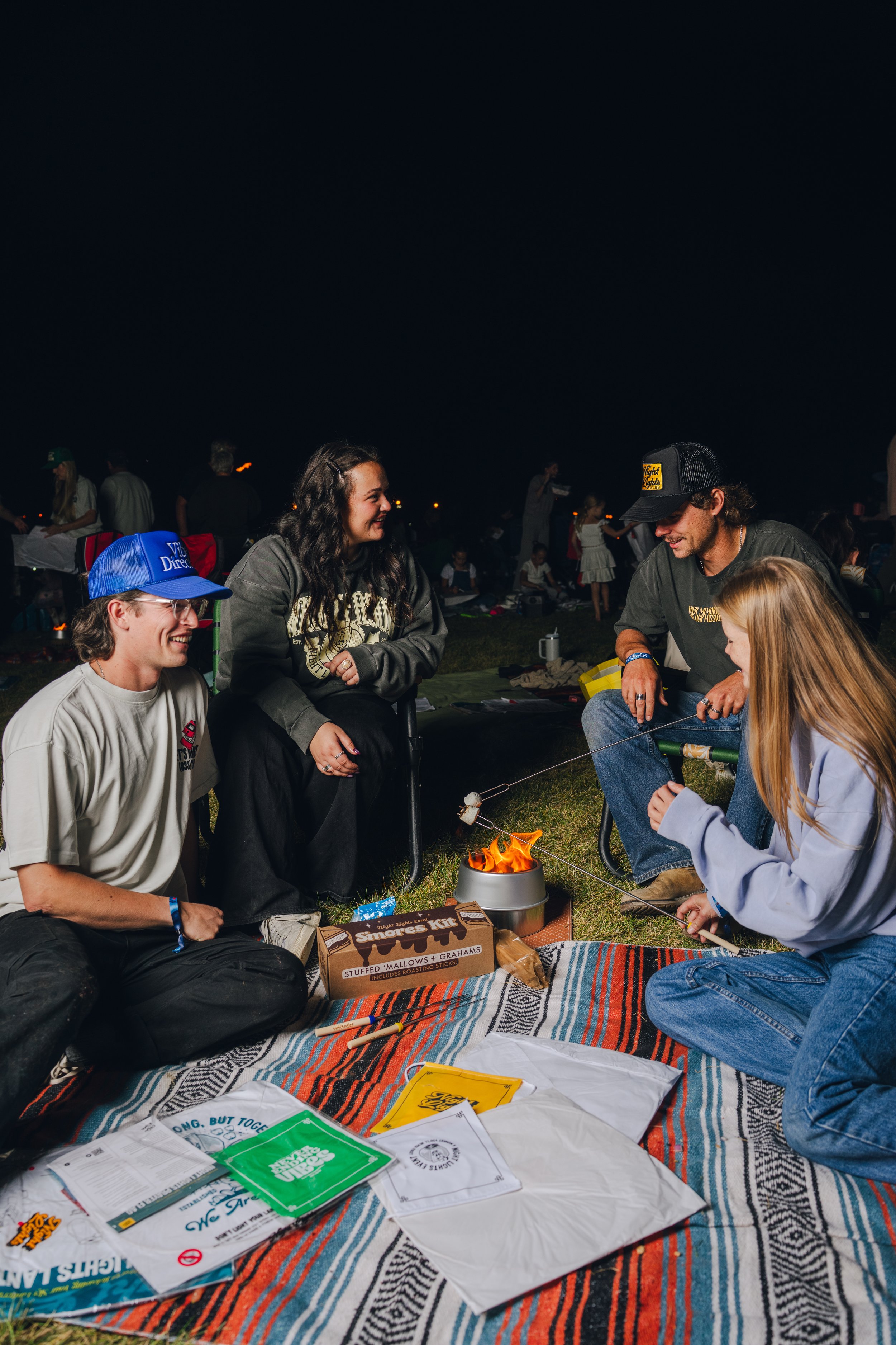 A group of people gathered outdoors at night, sitting on a colorful blanket, with a small fire in the middle. They are smiling and engaged in a conversation. There are various papers and snacks on the blanket.
