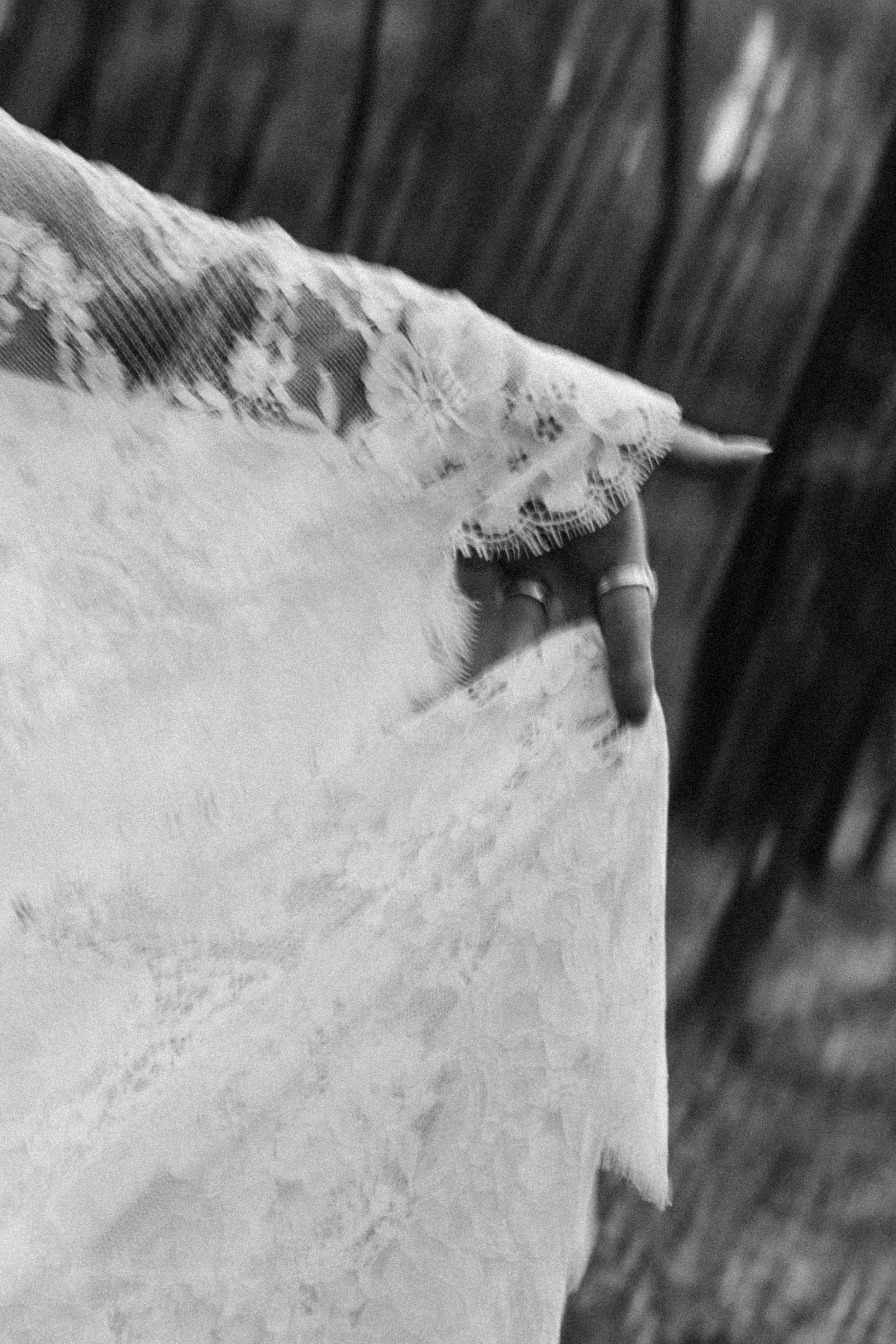 Close-up black and white photo of a person's finger touching a lace fabric on a surface, with wooden planks in the background.