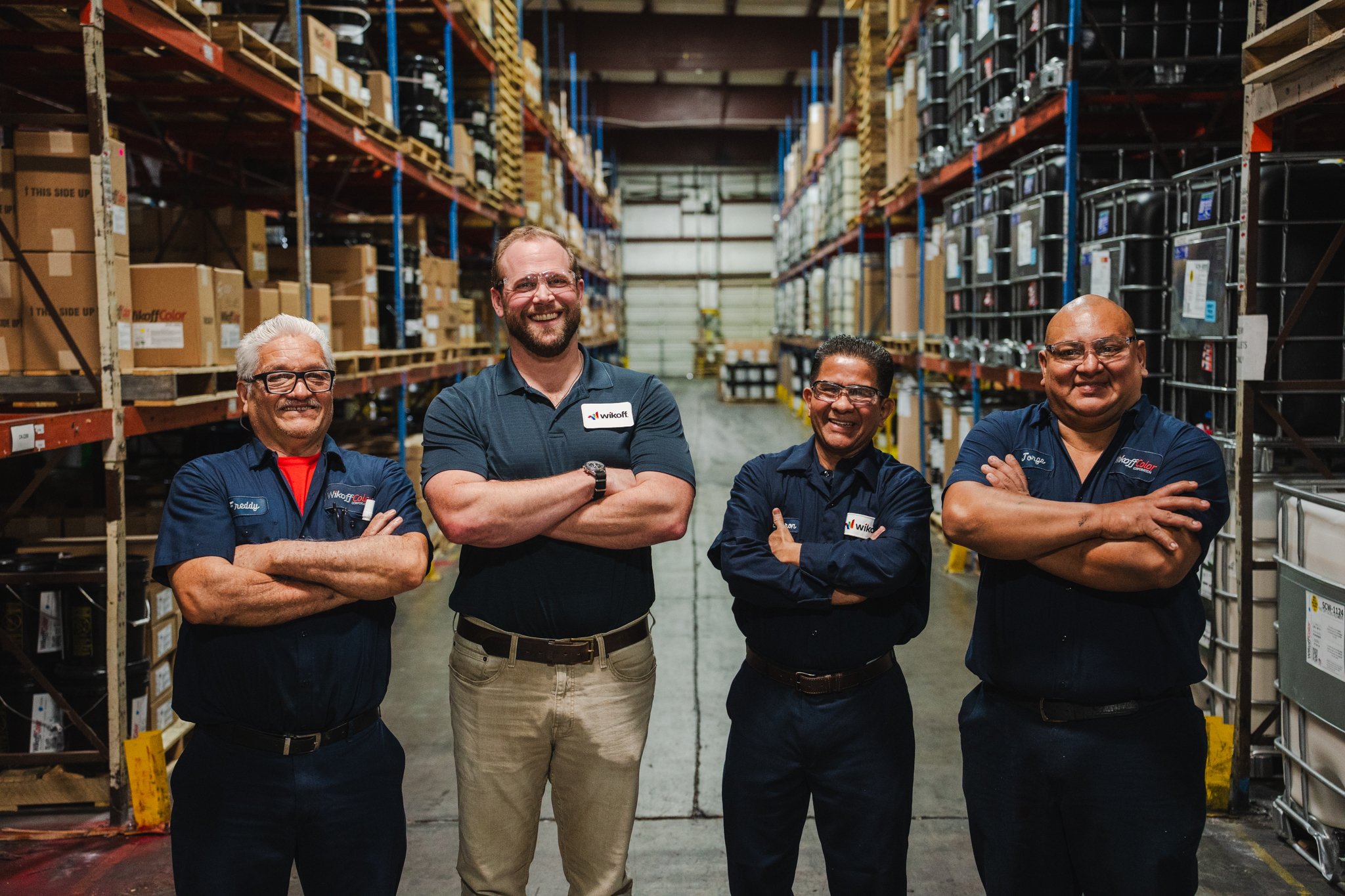 Four smiling warehouse employees standing with arms crossed in aisle surrounded by shelves filled with boxes and hardware.