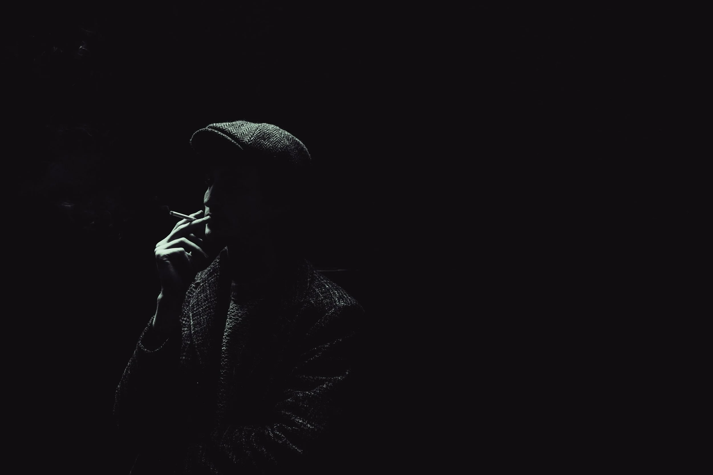 A person wearing a hat and coat, smoking a cigarette in low light with a dark background.
