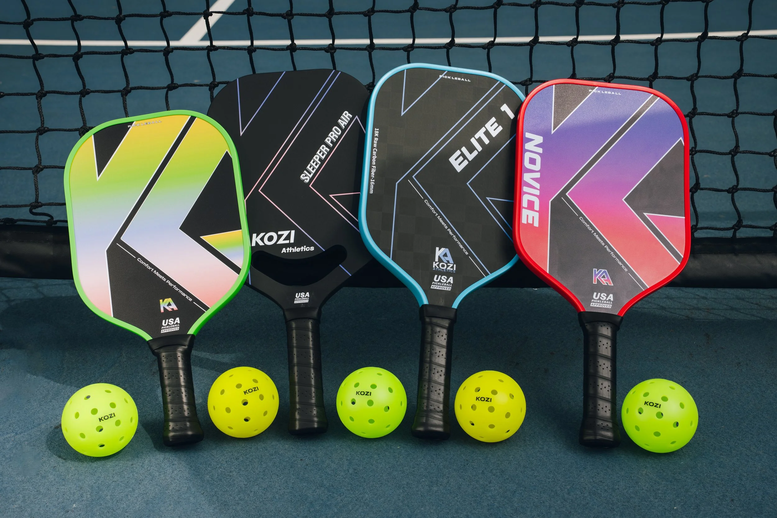 Four pickleball paddles in different colors (green, black, blue, red) and five yellow perforated pickleballs arranged on a blue surface in front of a black net.