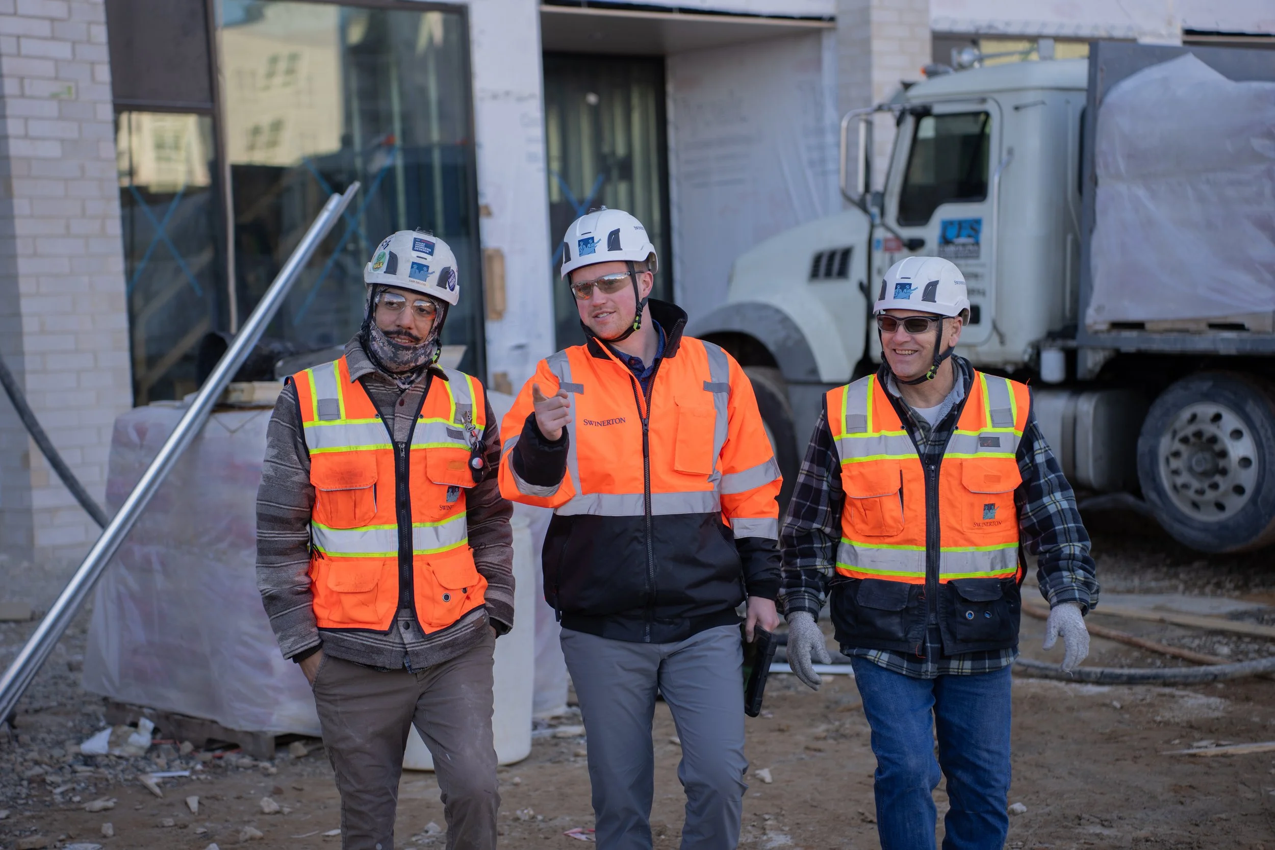Three construction workers in safety vests and helmets walking and talking on a construction site with a building and a truck in the background.