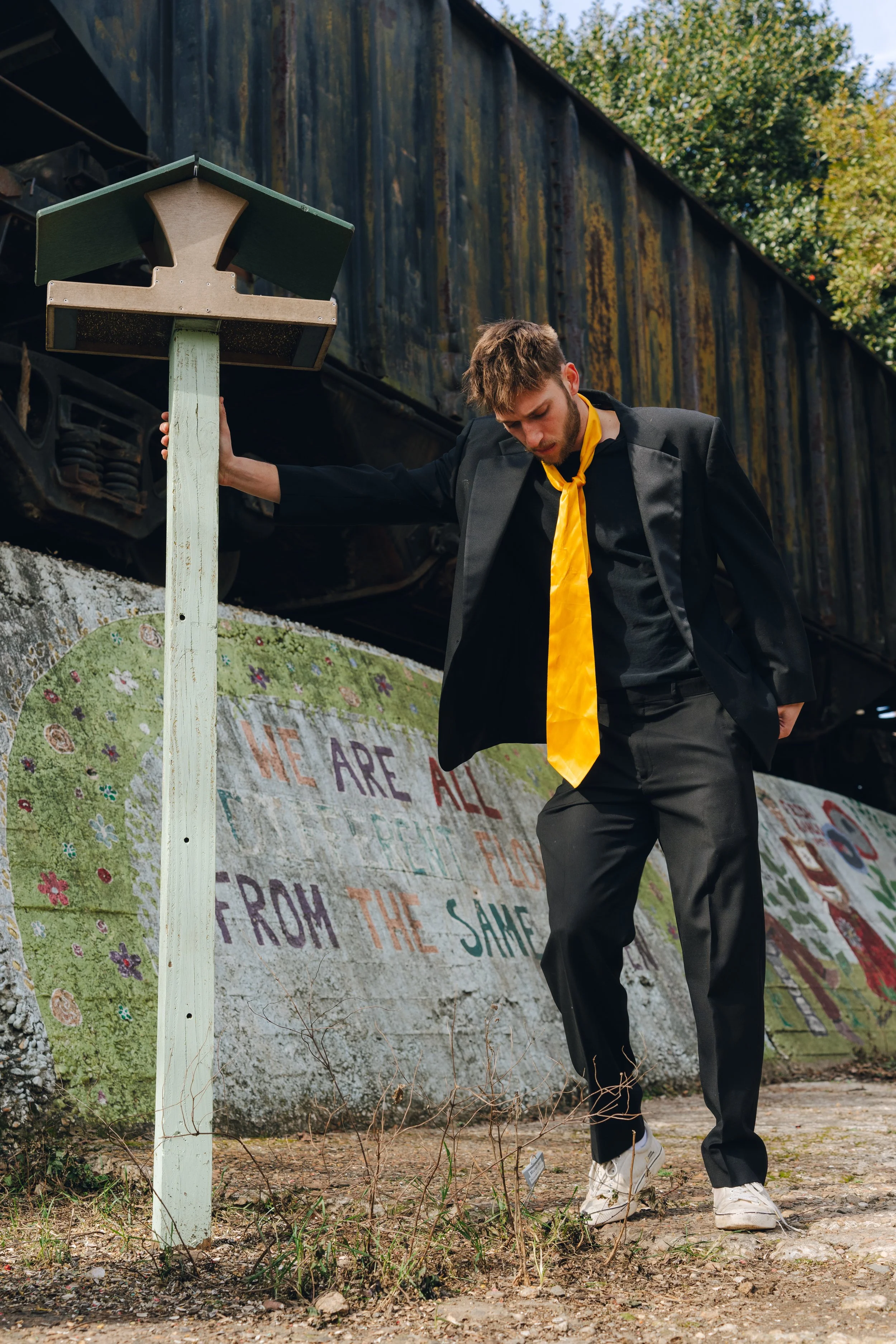 A young man in a black suit and yellow scarf standing outdoors, looking down, near a lamppost and a colorful mural painted on a concrete wall with plants and trees in the background.