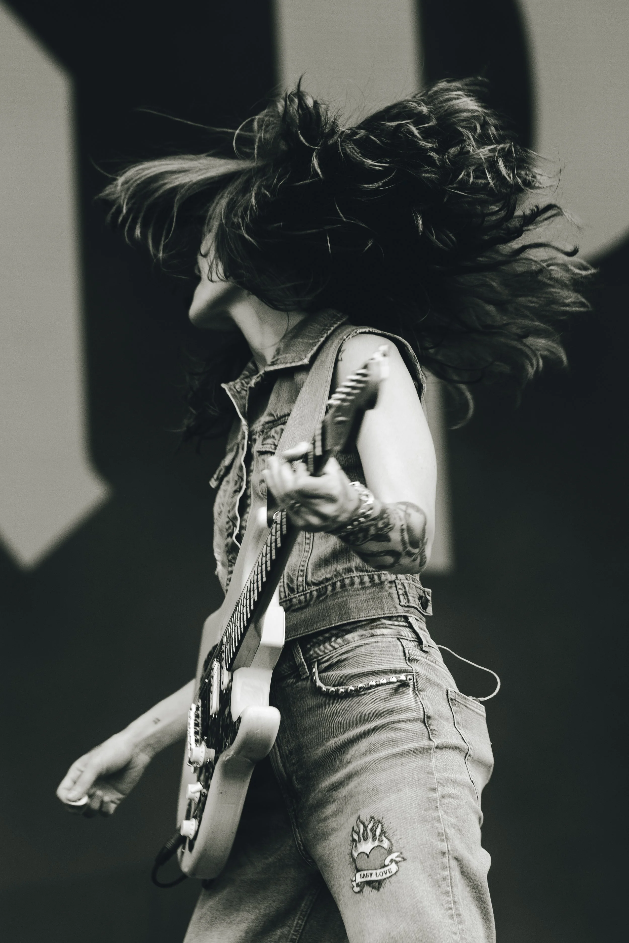 A person with long, curly hair playing an electric guitar. The person is wearing a sleeveless denim vest and jeans with a tattoo on their thigh that reads 'Early Love' with a heart and flames. The image is in black and white.