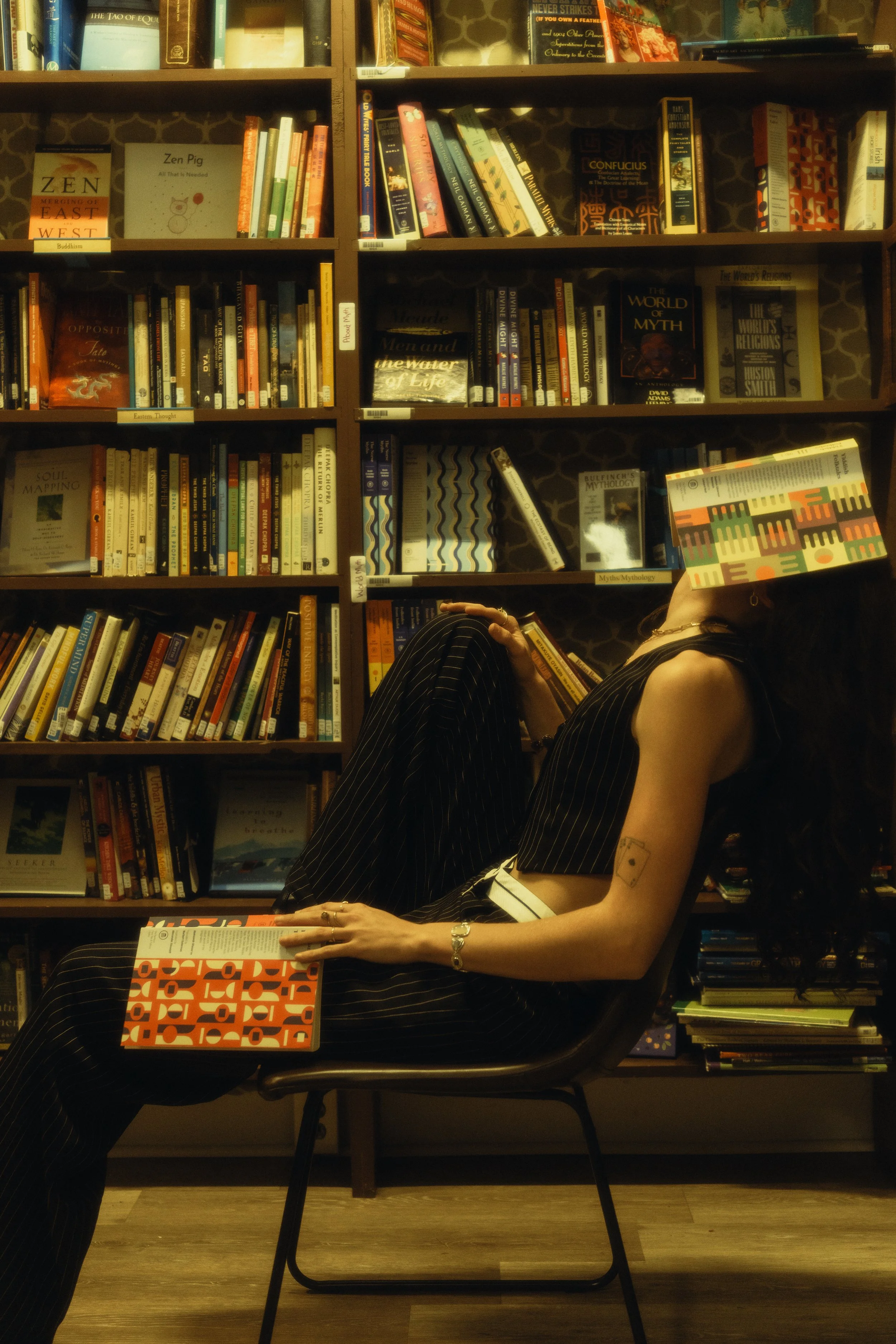 A person sitting in a library holding a book, with a bookshelf filled with various books behind them. The person is wearing a striped black outfit and has tattoos on their arm, and the photo is taken in a reflective, slightly tilted angle.