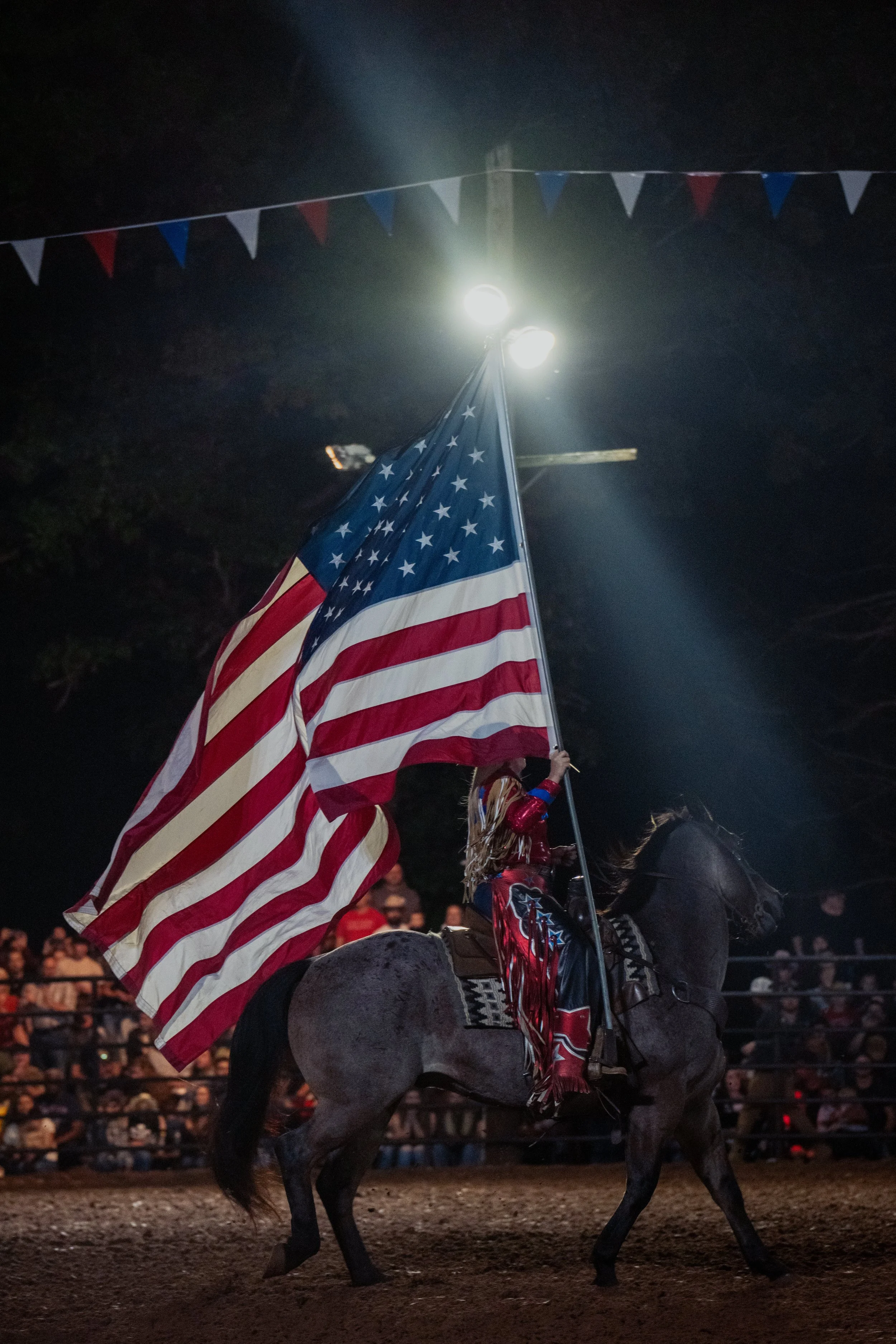 A person riding a horse at night during a rodeo, holding an American flag, with parade decorations overhead and a crowd in the background.