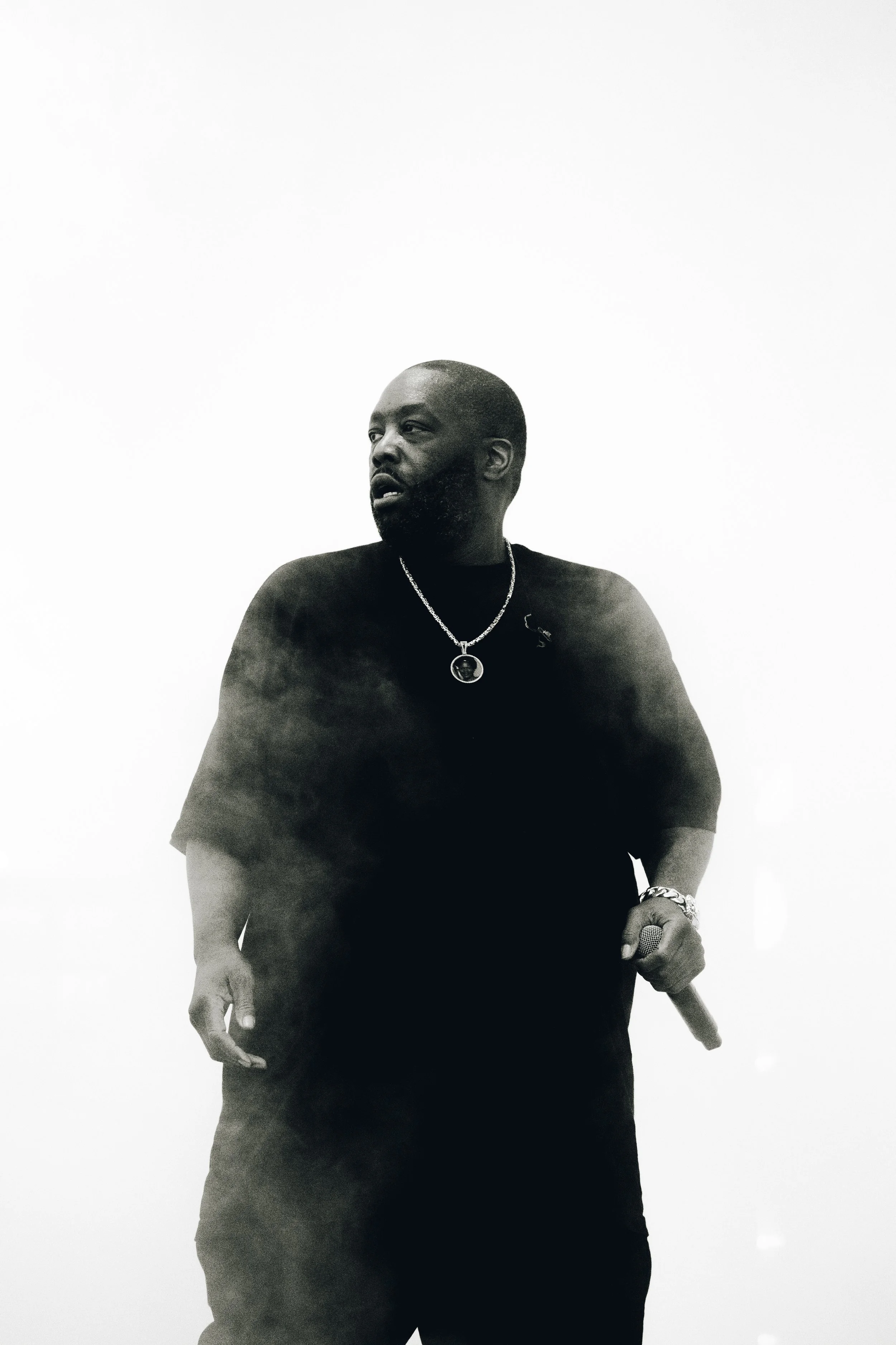 A man with a beard and short hair, wearing a black t-shirt, silver chain necklace, and bracelet, holds a microphone, standing against a plain white background.