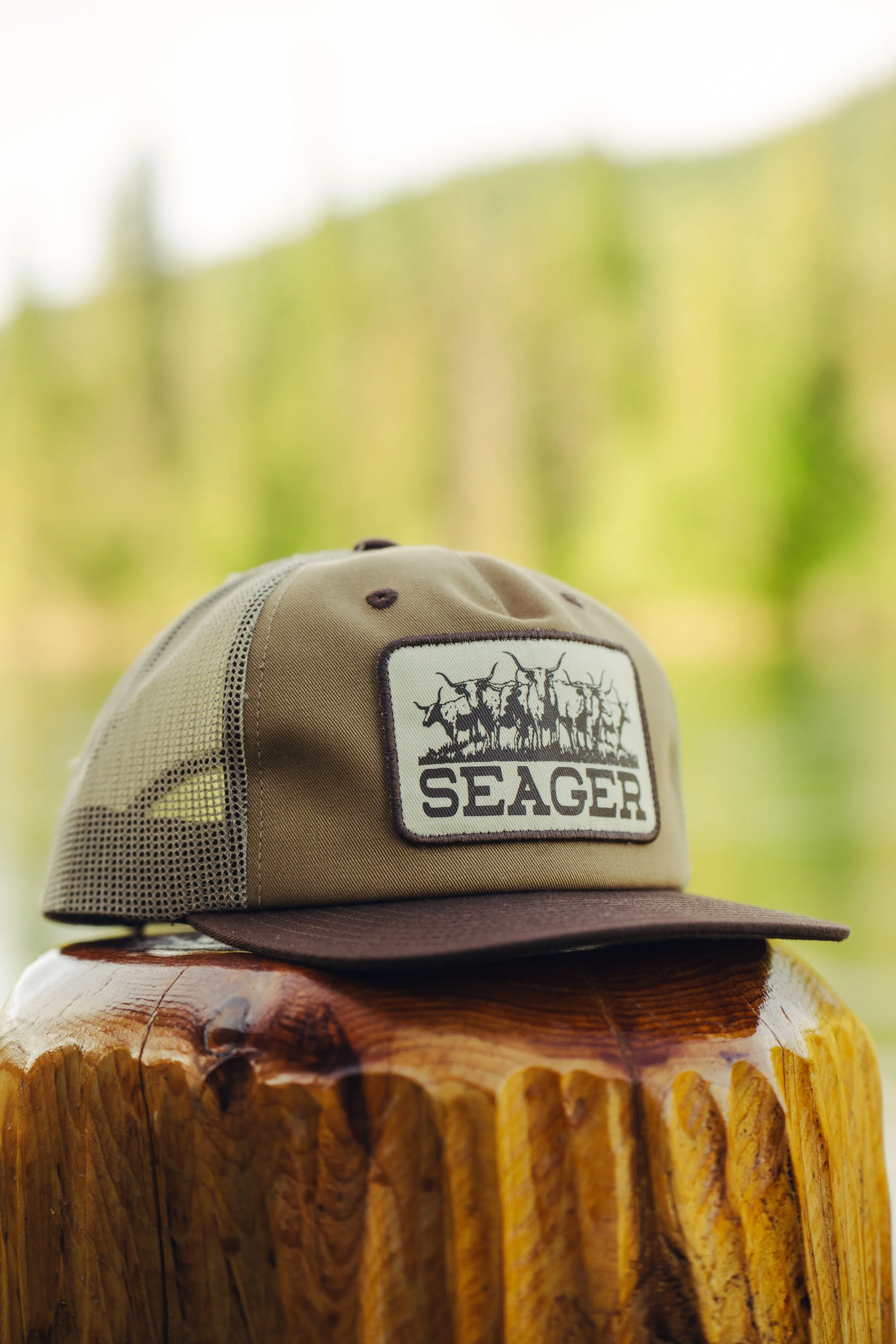A beige and brown baseball cap with a 'Seager' patch featuring a graphic of a hunting scene, placed on a wooden surface with a blurred natural green background.