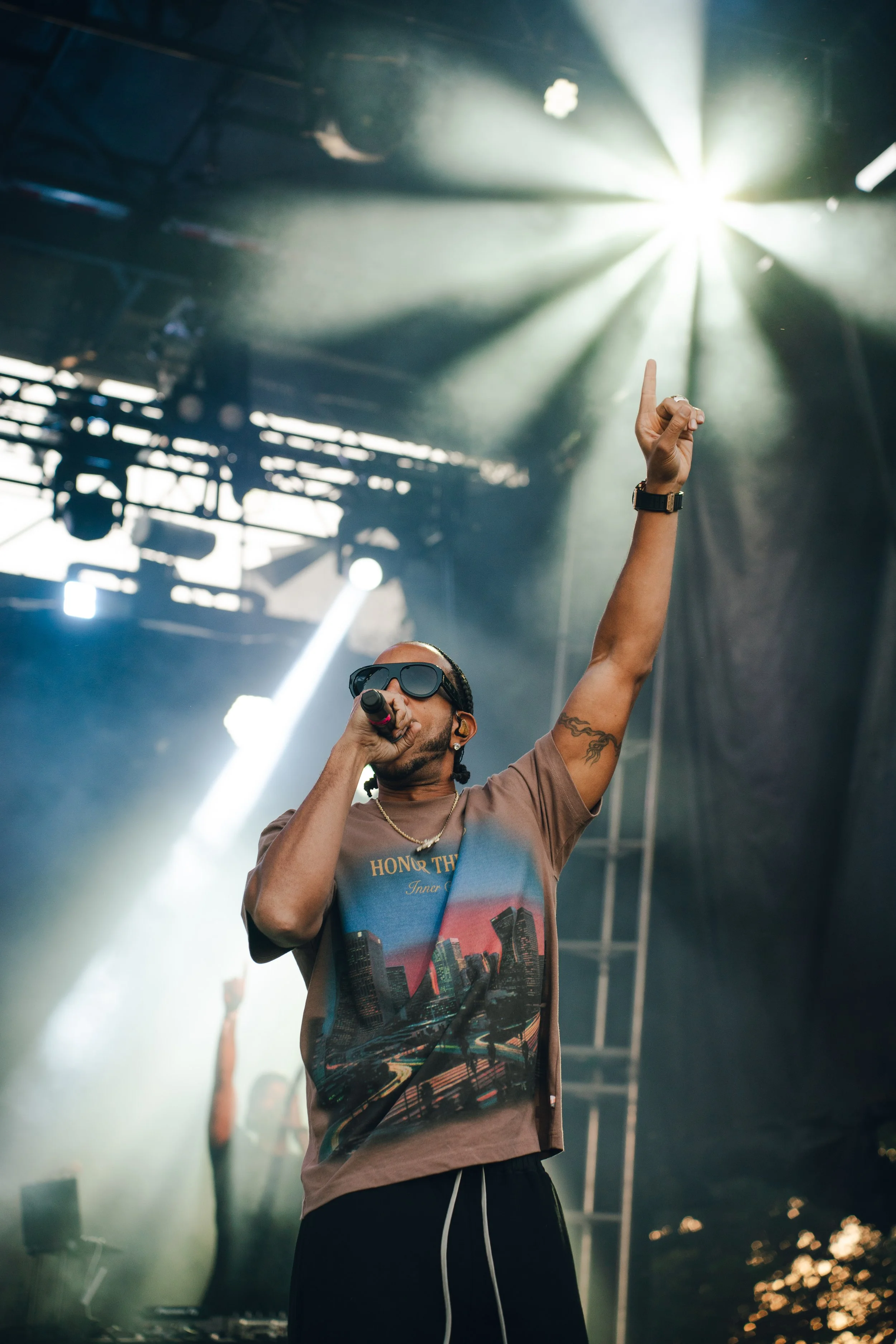 A male performer on stage wearing sunglasses and a graphic T-shirt, holding a microphone, with one arm raised, during a concert with stage lights and fog.
