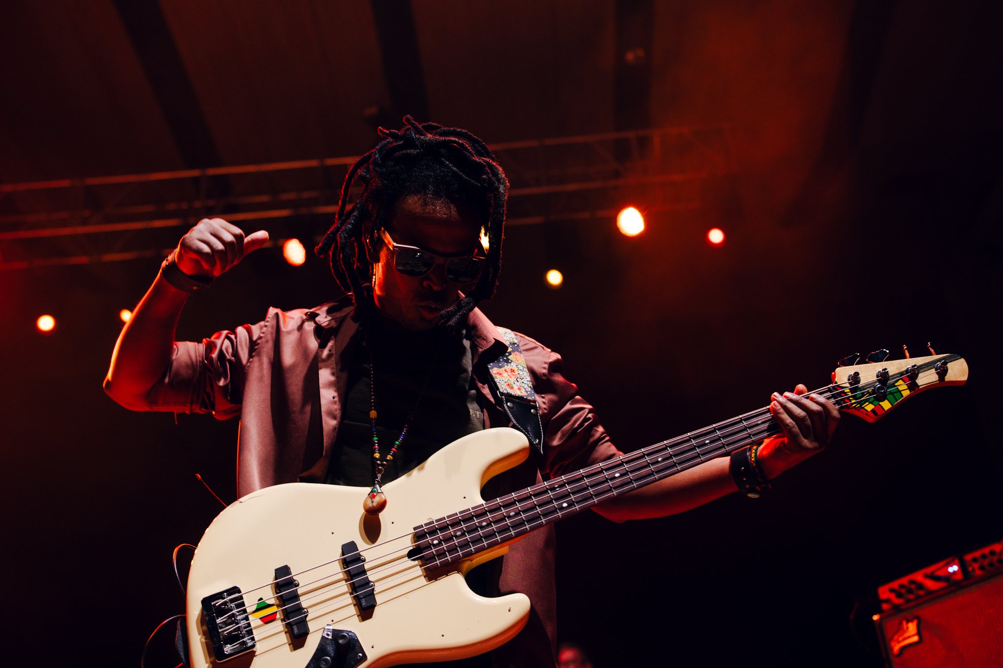 A musician with dreadlocks, sunglasses, and jewelry playing an electric bass guitar on stage, illuminated by orange stage lights.