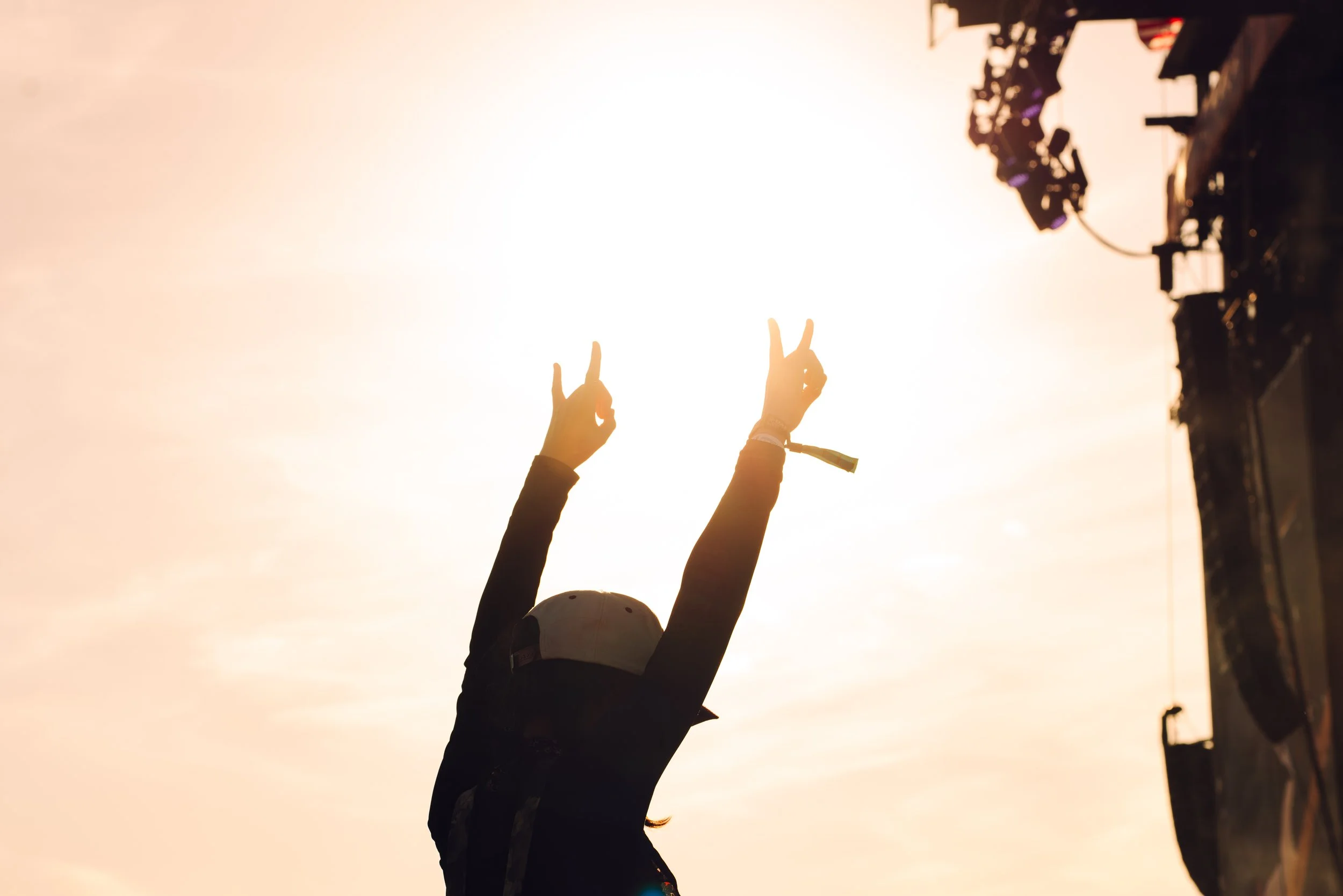 Person making a peace sign with both hands at sunset, with stage equipment visible on the right.