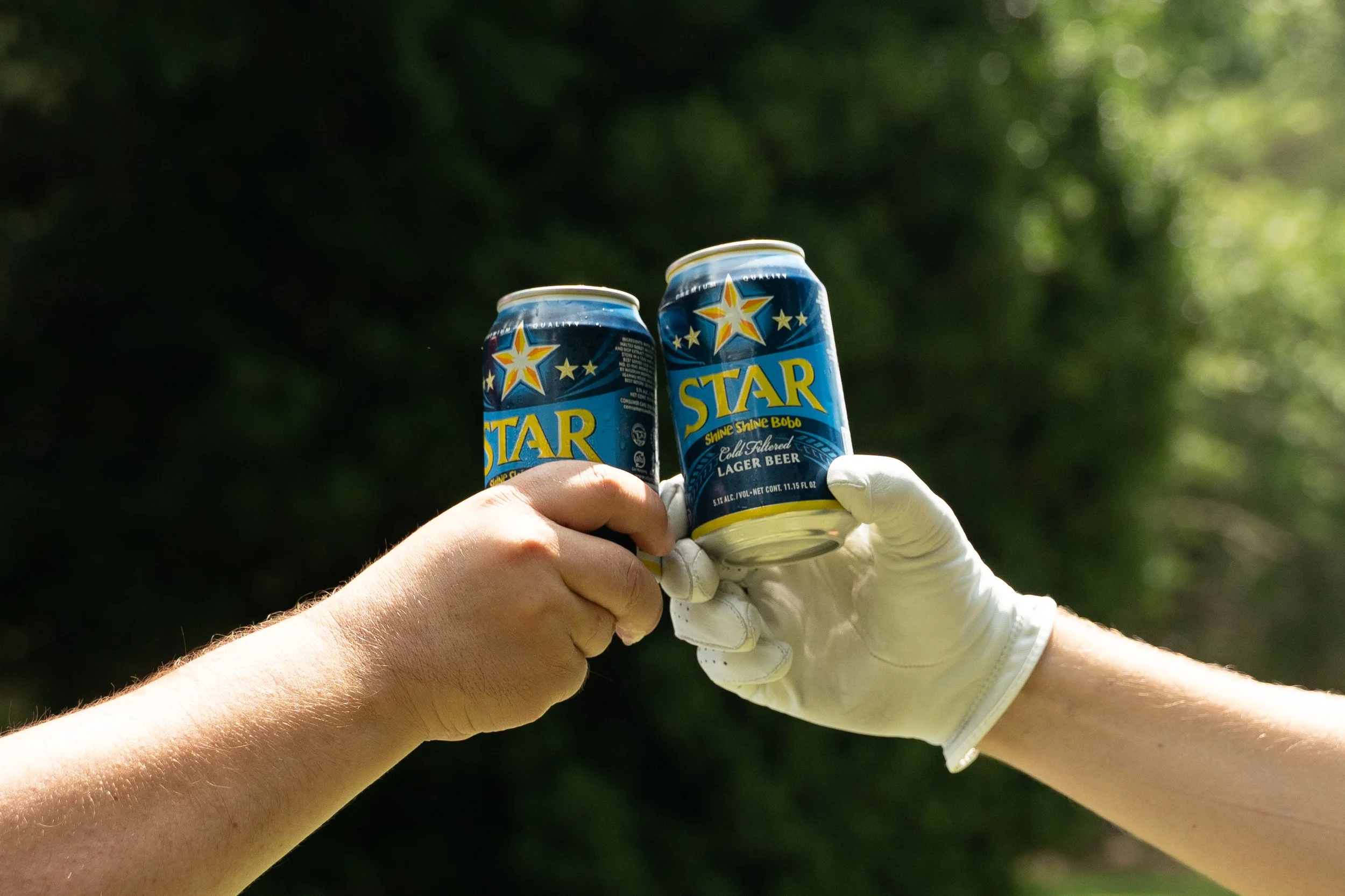 Two people holding cans of Star Shine Shine Bobo beer outdoors with blurred green foliage in the background.