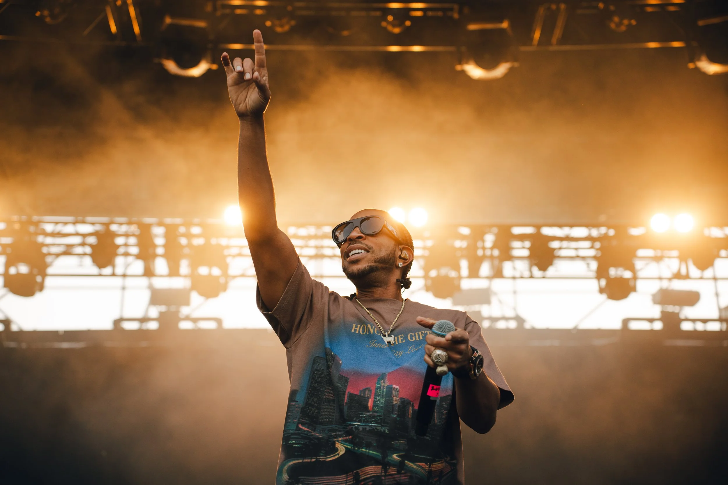 Musician performing on stage with lighting behind him, wearing sunglasses, a brown t-shirt, and holding a microphone, raising his hand.