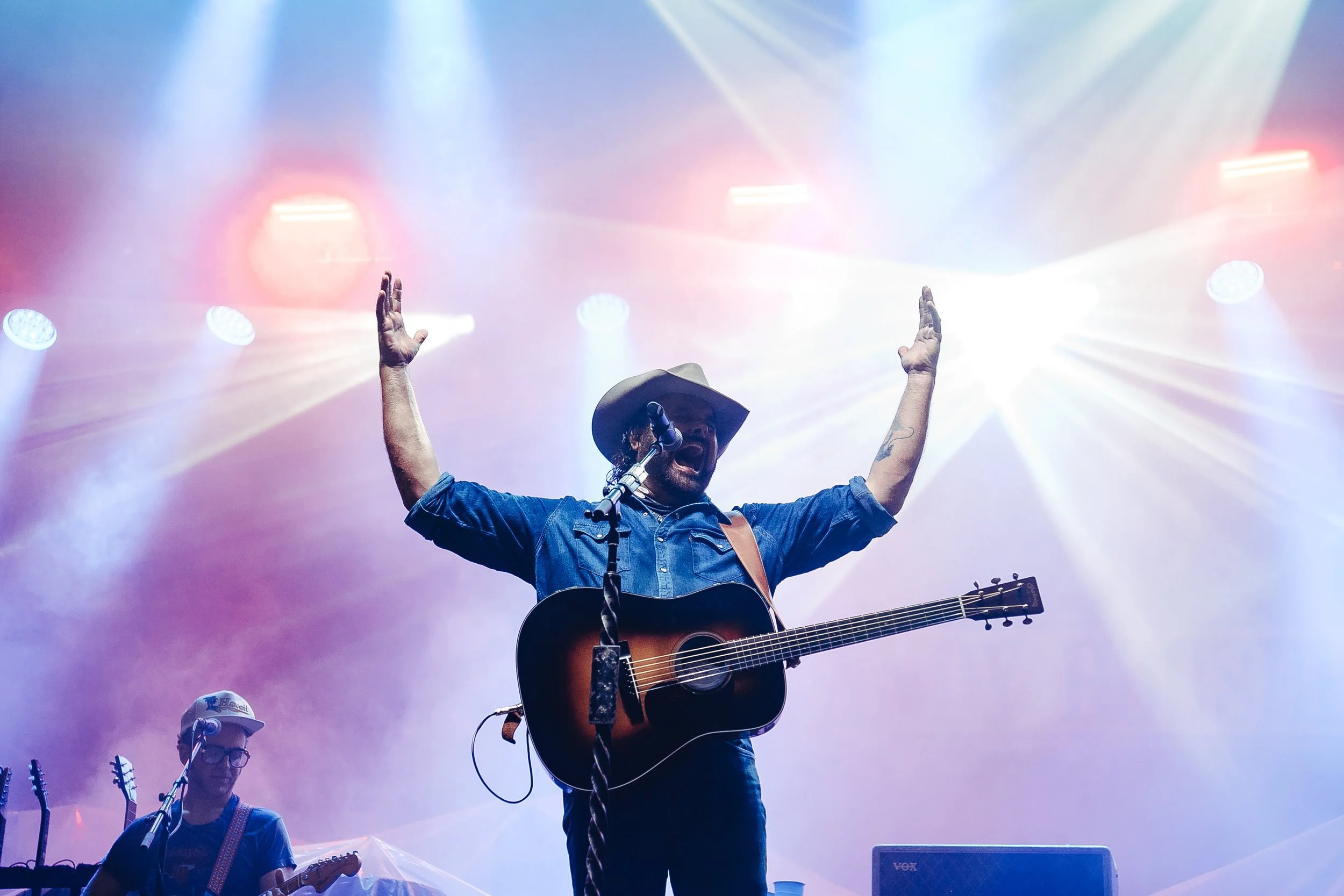 A male musician wearing a hat and playing an acoustic guitar on stage, with his arms raised in the air, illuminated by colorful bright stage lights at a concert.