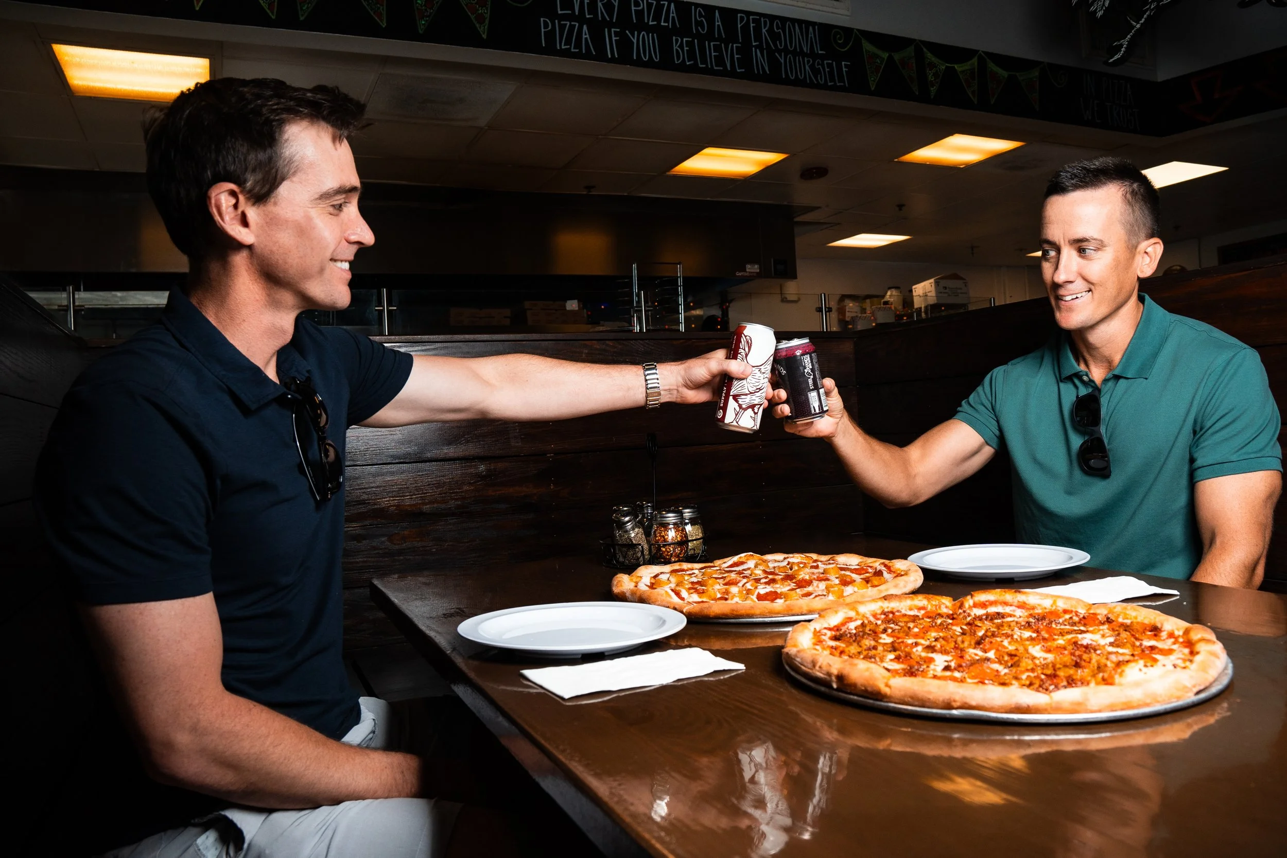 Two men sharing a toast with canned drinks in a pizza restaurant, with pizzas on the table and a motivational quote on the wall in the background.
