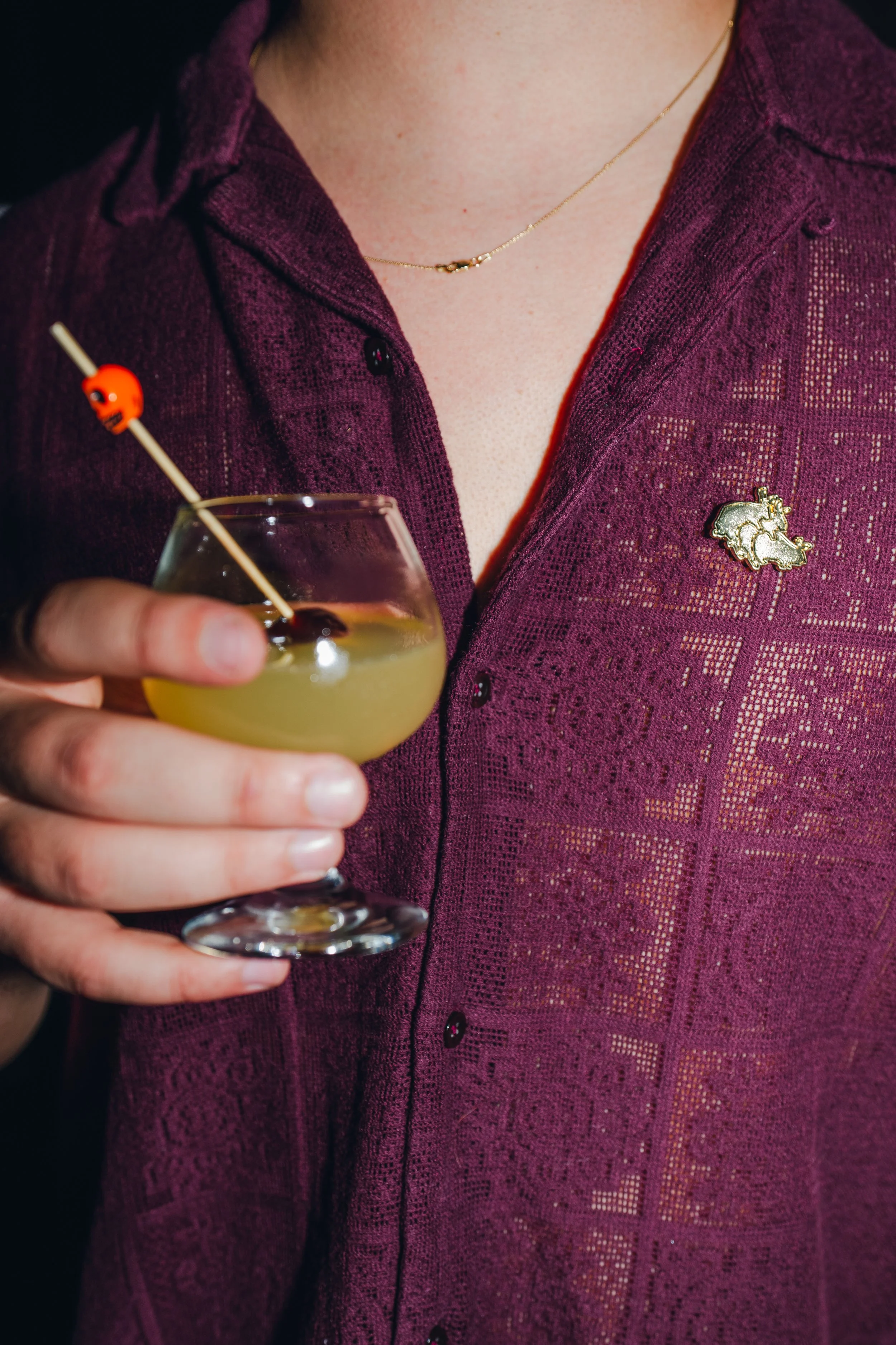 Person wearing a dark purple patterned shirt with a star-shaped pin and a delicate gold necklace, holding a cocktail glass with a yellow drink and a small skewer with an orange skull decoration.