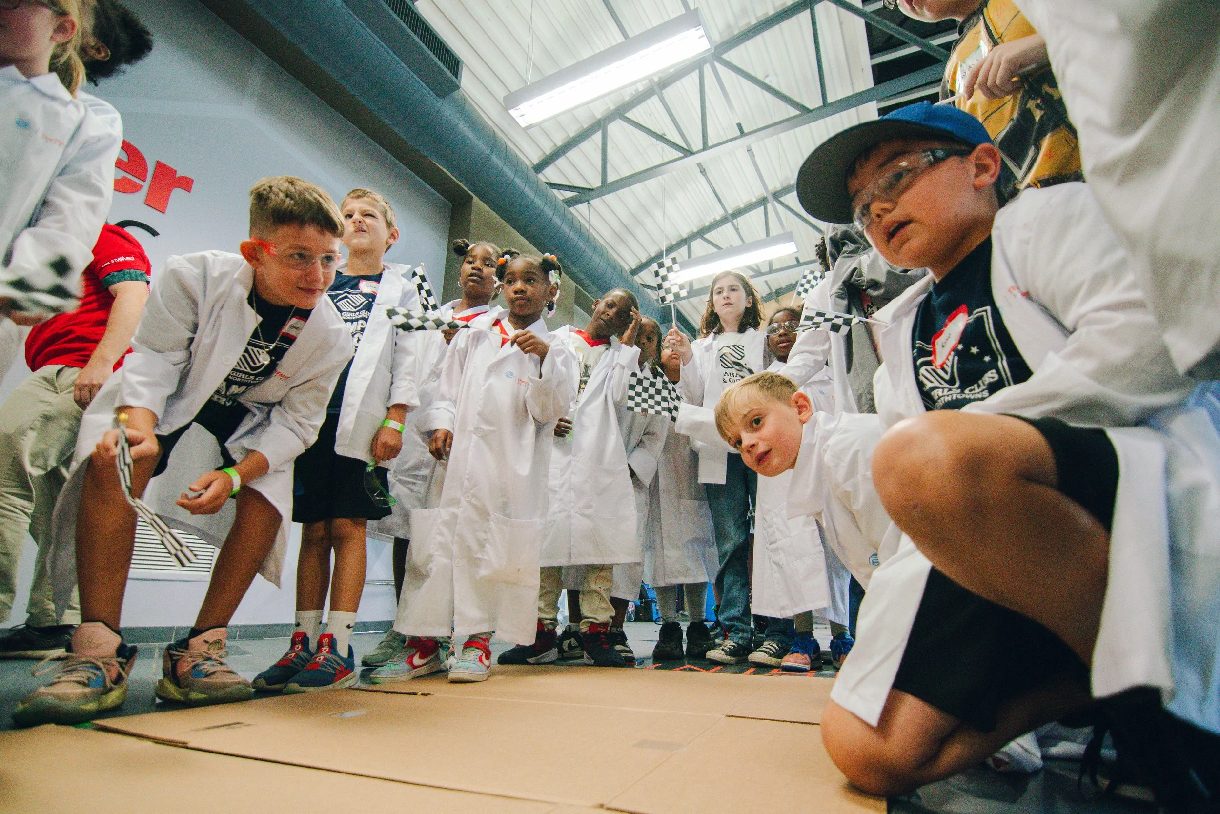 Children gathered in a line, some wearing lab coats and safety goggles, participating in a science or STEM activity in a large indoor space with a high, metal ceiling.