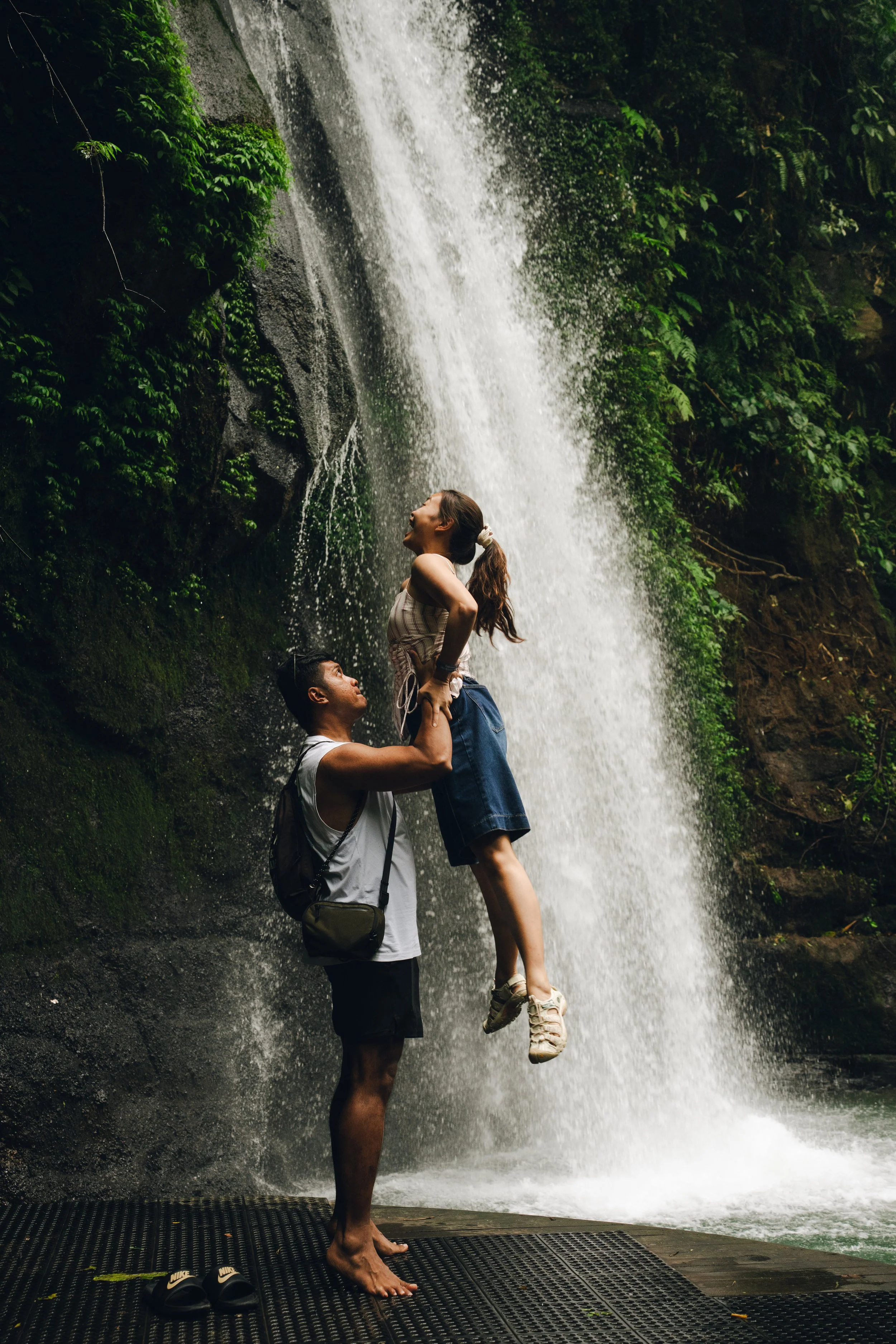 A man holding a woman in front of a waterfall on a wooden platform, with a pair of black Nike slide sandals on the platform in the foreground.