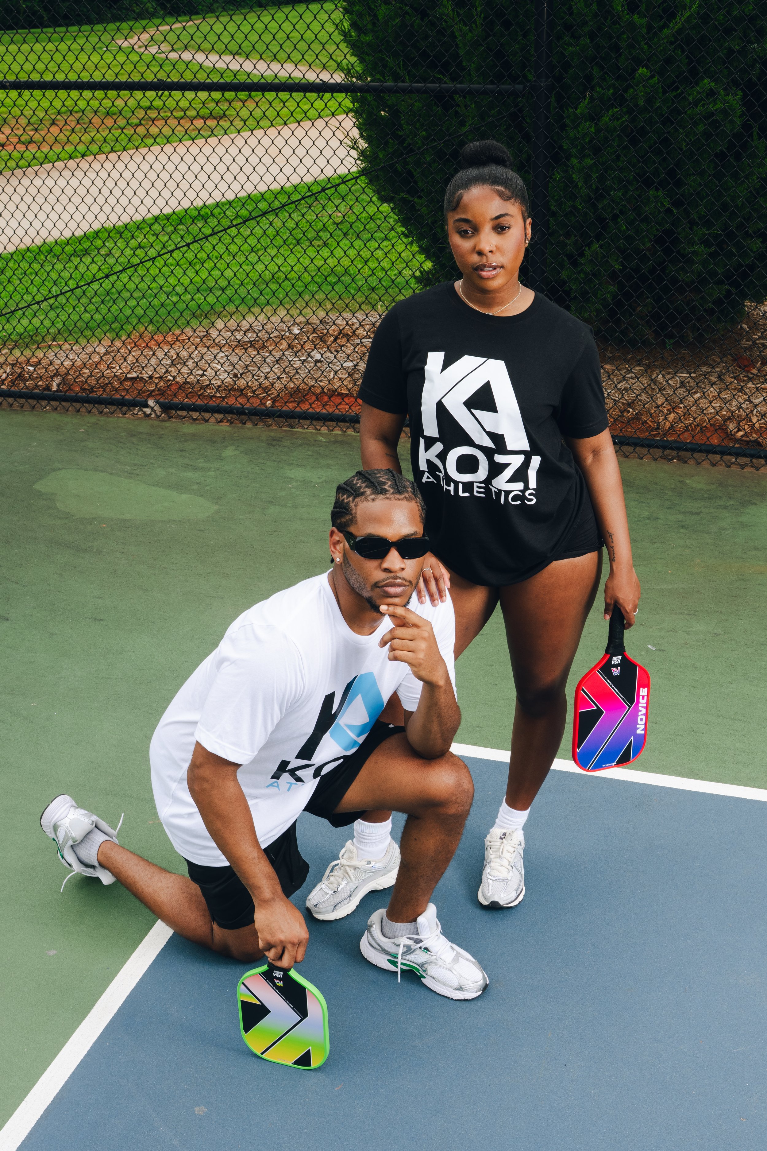 Two young adults on a tennis court, one kneeling and the other standing, both holding colorful paddle tennis rackets, with a fence and green trees in the background.