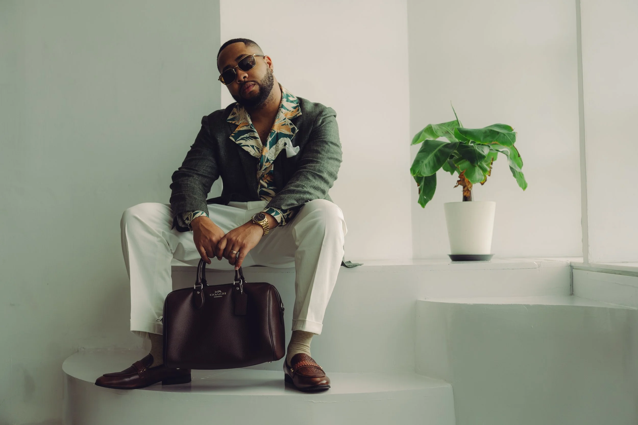 A man with a beard and dark sunglasses sits on a white rounded platform, wearing a patterned shirt under a dark blazer, white pants, and brown dress shoes. He holds a dark brown Coach handbag, with a large potted plant with broad green leaves beside 