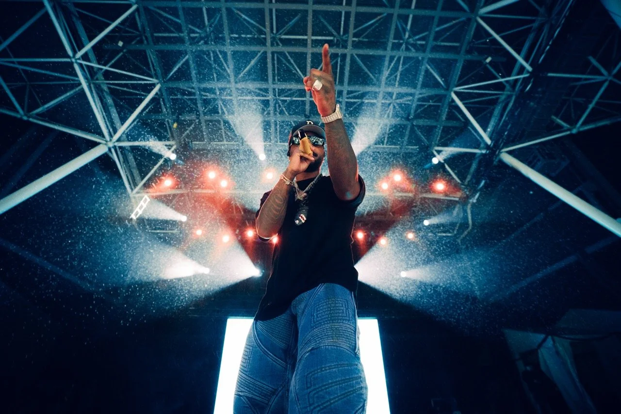 A male performer on stage at a concert or festival, holding a microphone, with bright stage lights and a large LED screen behind him.