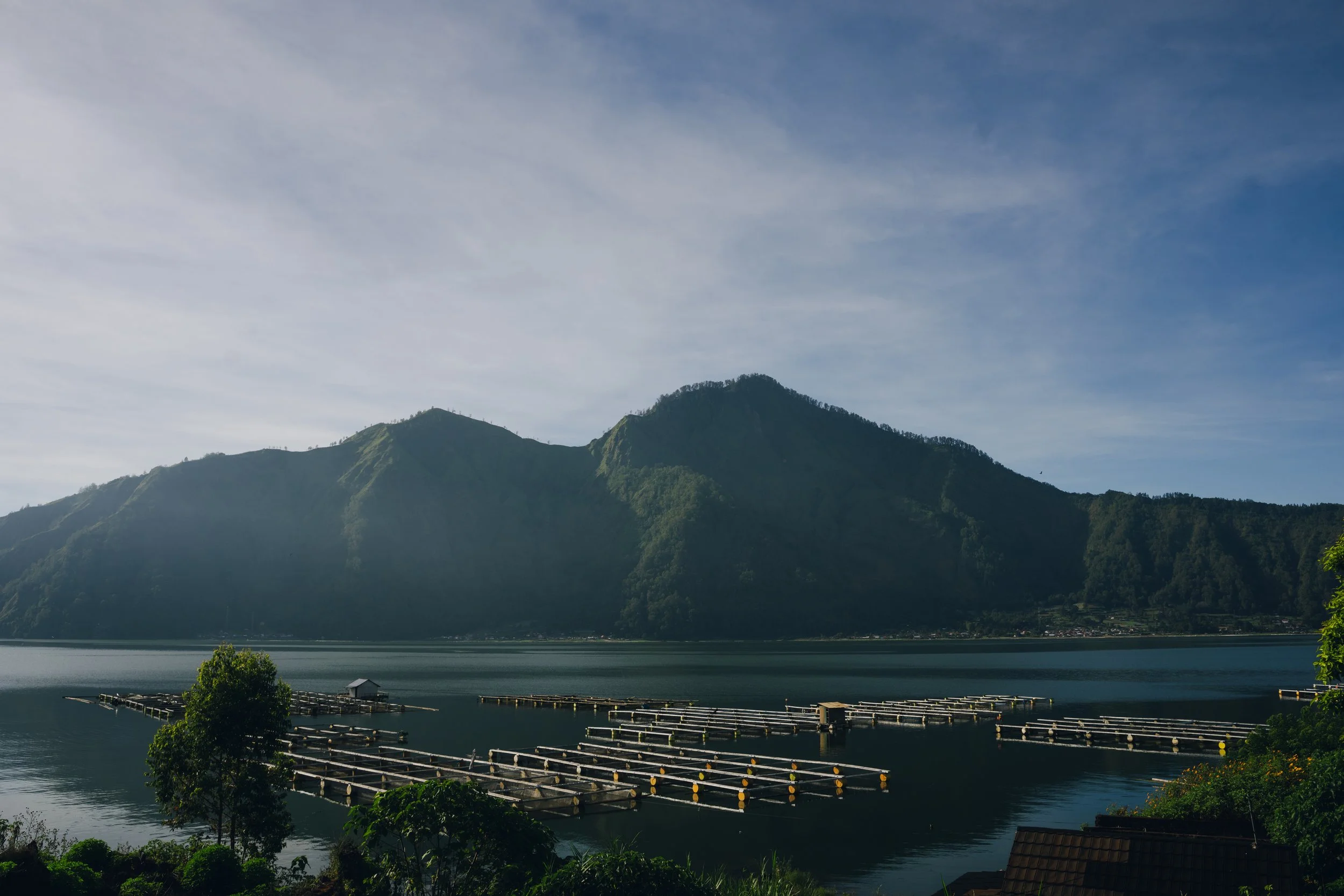 A scenic view of a mountain range with a lake in the foreground, dock structures on the lake, and trees along the shoreline under a partly cloudy sky.