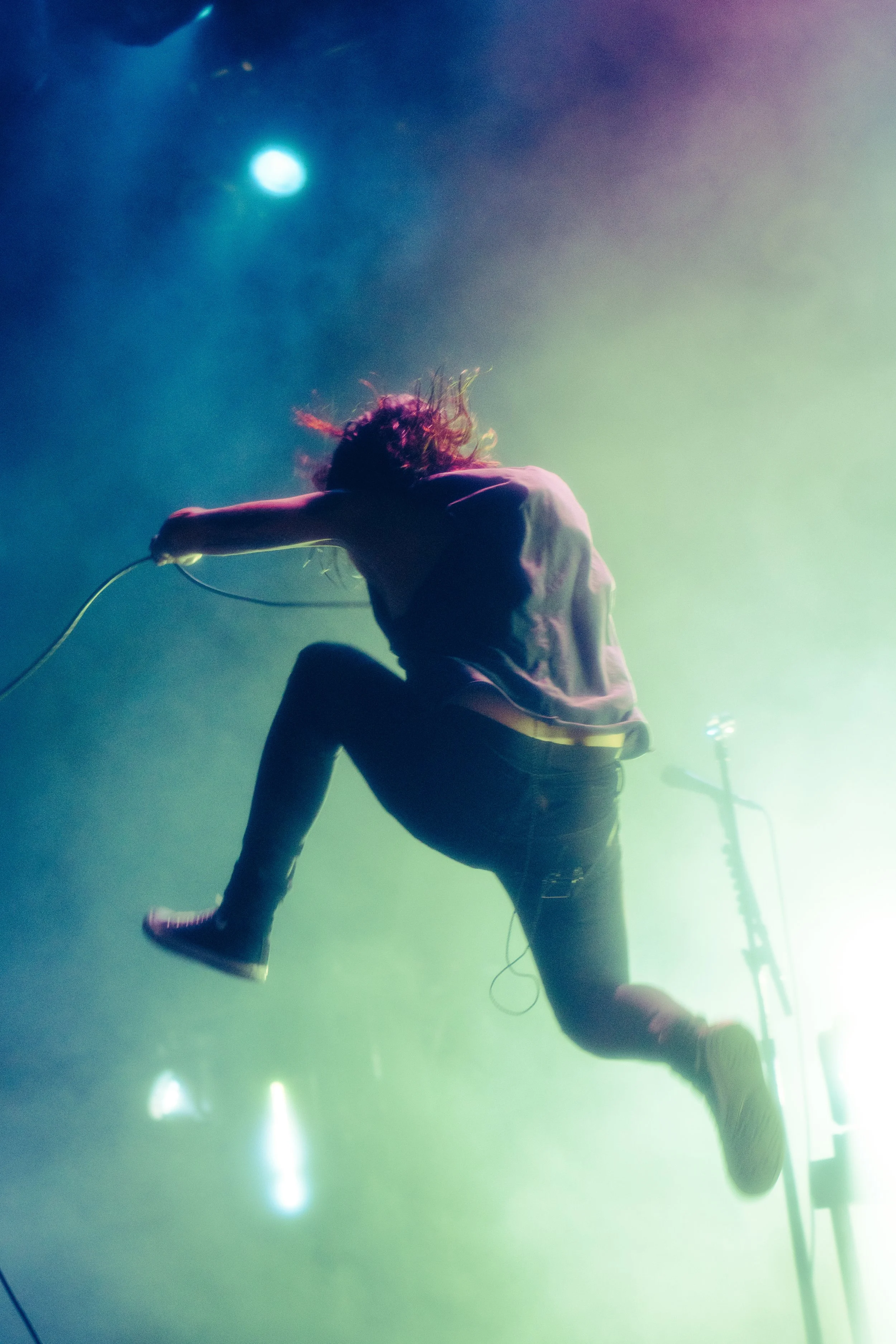 A performer in casual clothing, including sneakers and rolled-up jeans, is jumping or dancing on stage with a microphone in hand. The stage is illuminated with colorful lights and fog, creating an energetic atmosphere.
