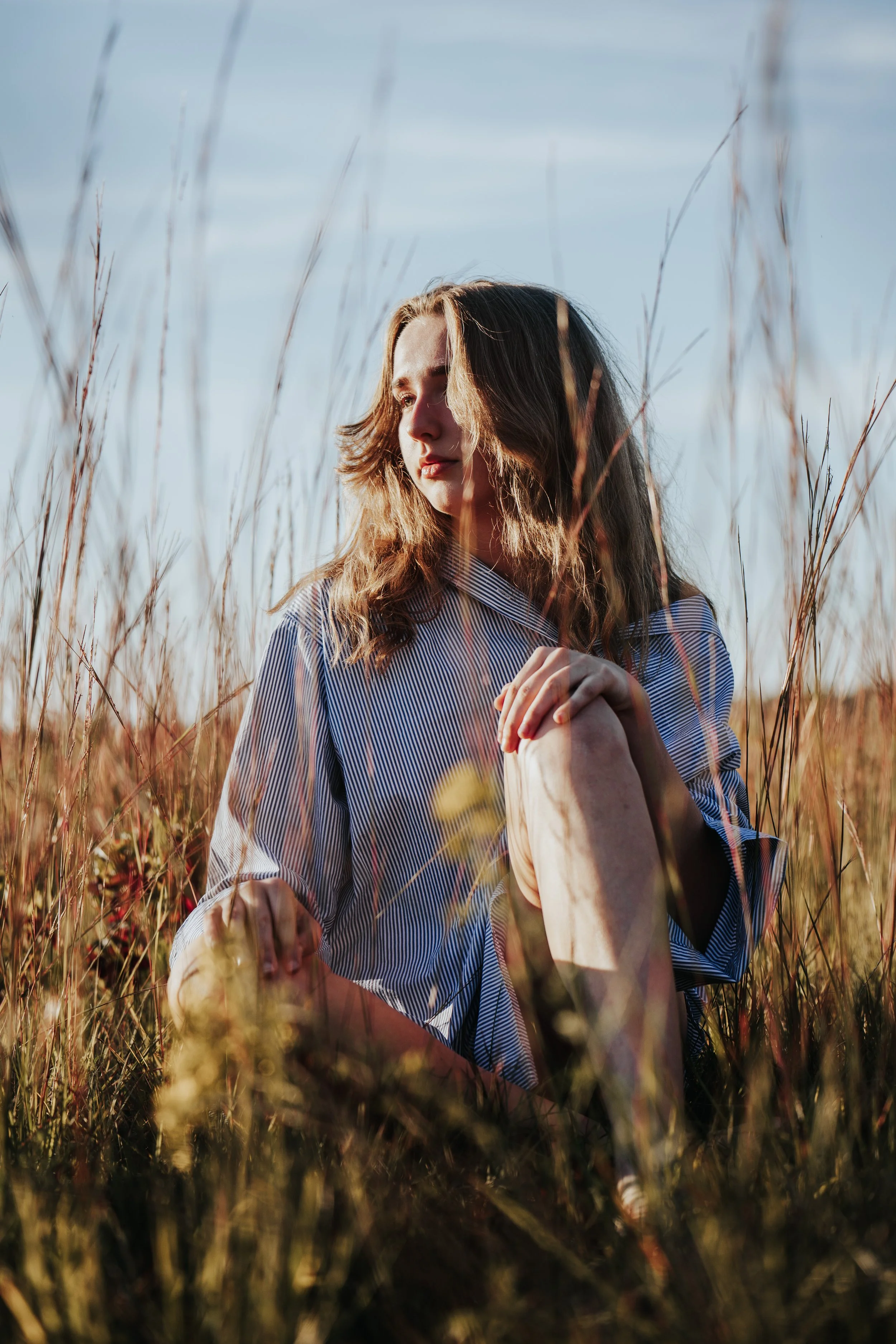 A woman with shoulder-length brown hair sitting in tall grass, wearing a striped shirt, with a calm expression and looking to the side, outdoors during daylight.