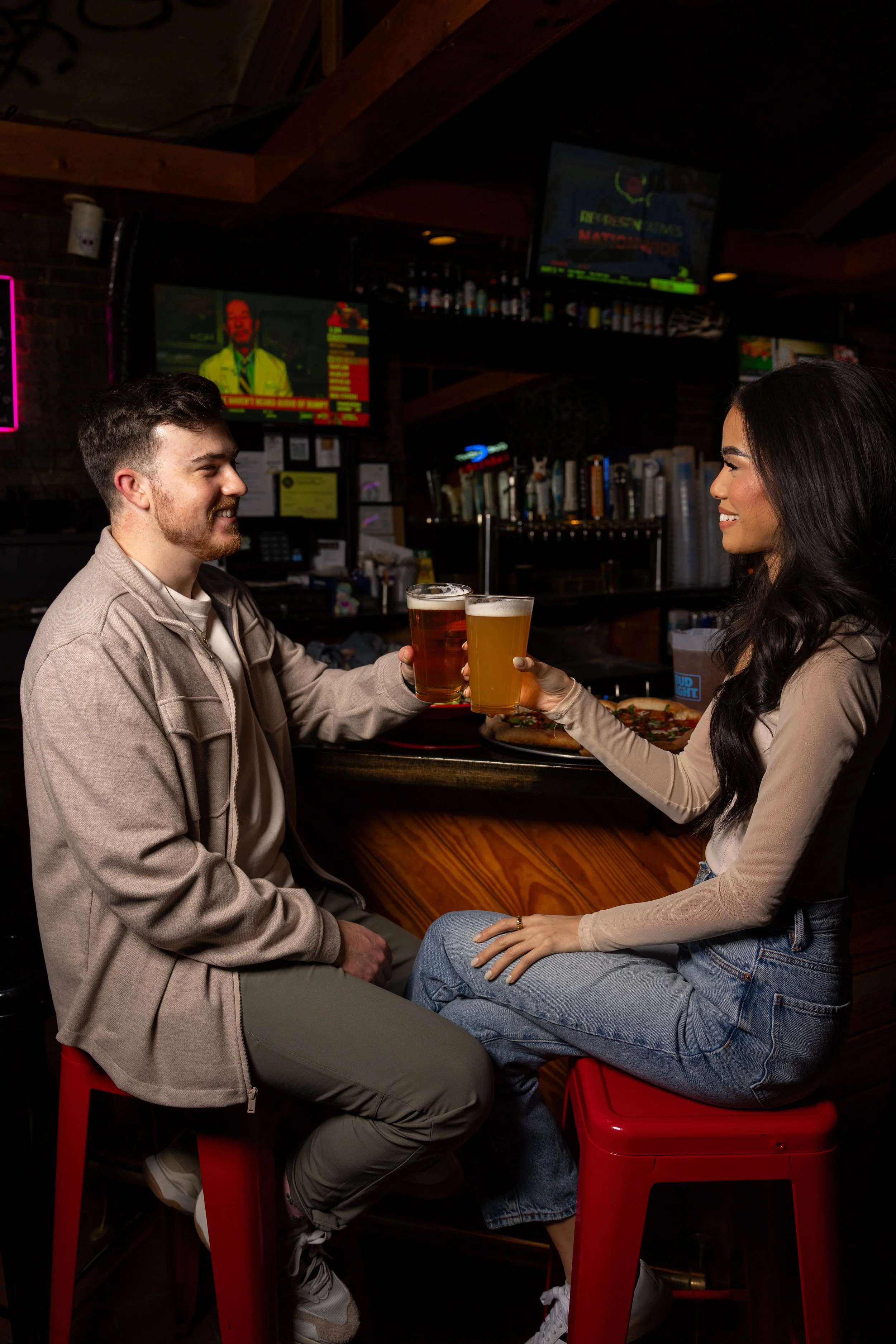 A young man and woman sitting at a bar, smiling and toasting each other with glasses of beer. The man has short brown hair and a beard, wearing a beige jacket and olive pants. The woman has long dark hair, wearing a beige top and blue jeans. There ar
