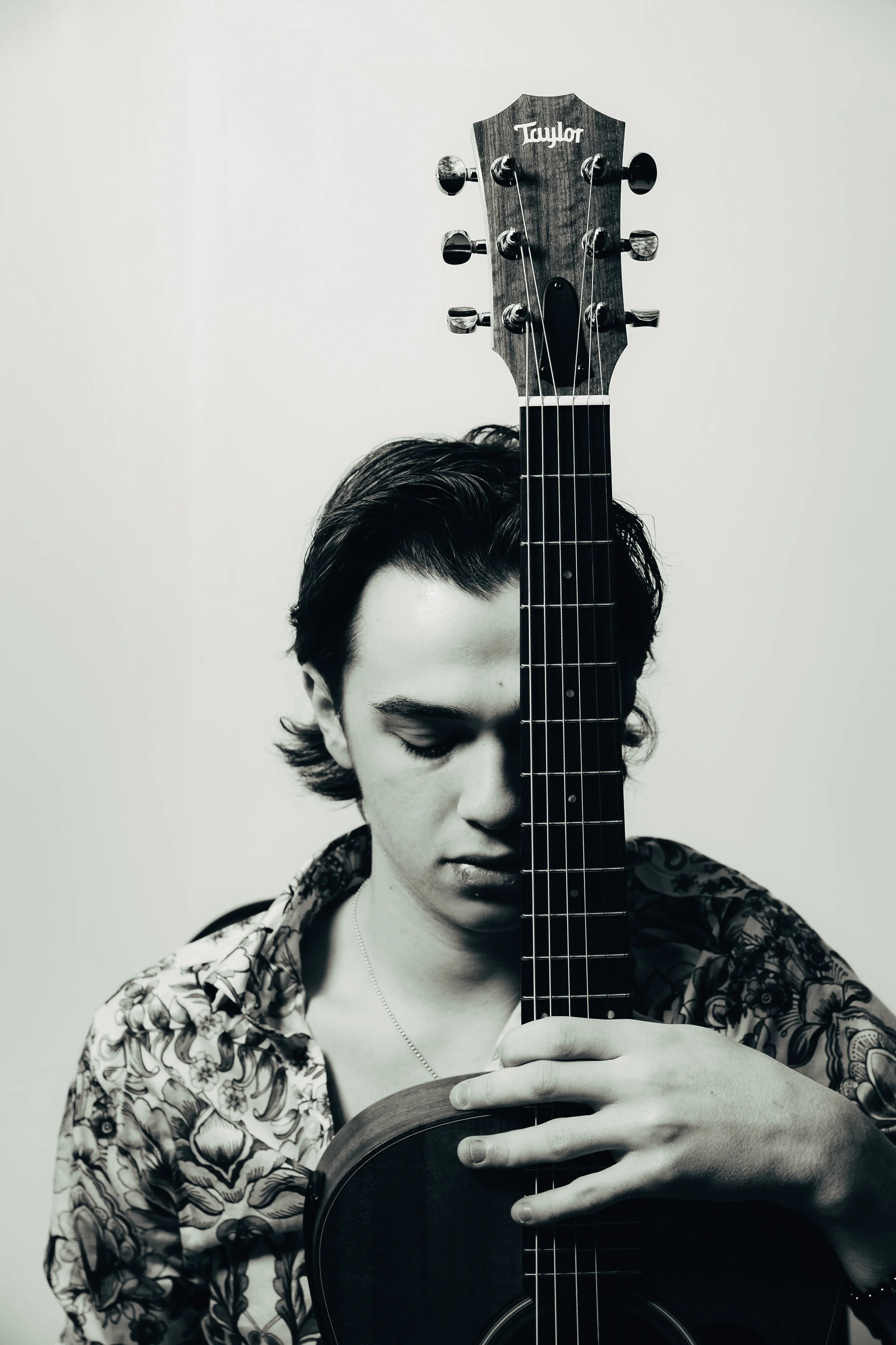 A black and white photo of a young man with dark hair, holding a guitar vertically in front of his face, with the headstock covering his eyes and forehead. The man is wearing a patterned shirt.