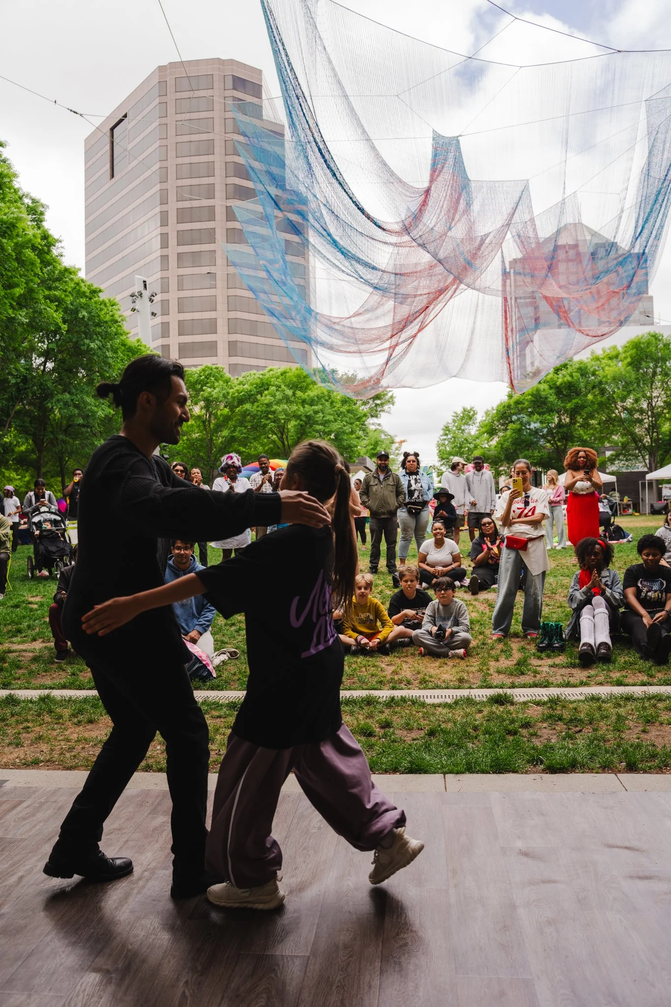 People dancing on a stage in front of a crowd in a park, with a large colorful art installation hanging overhead and tall city buildings in the background.