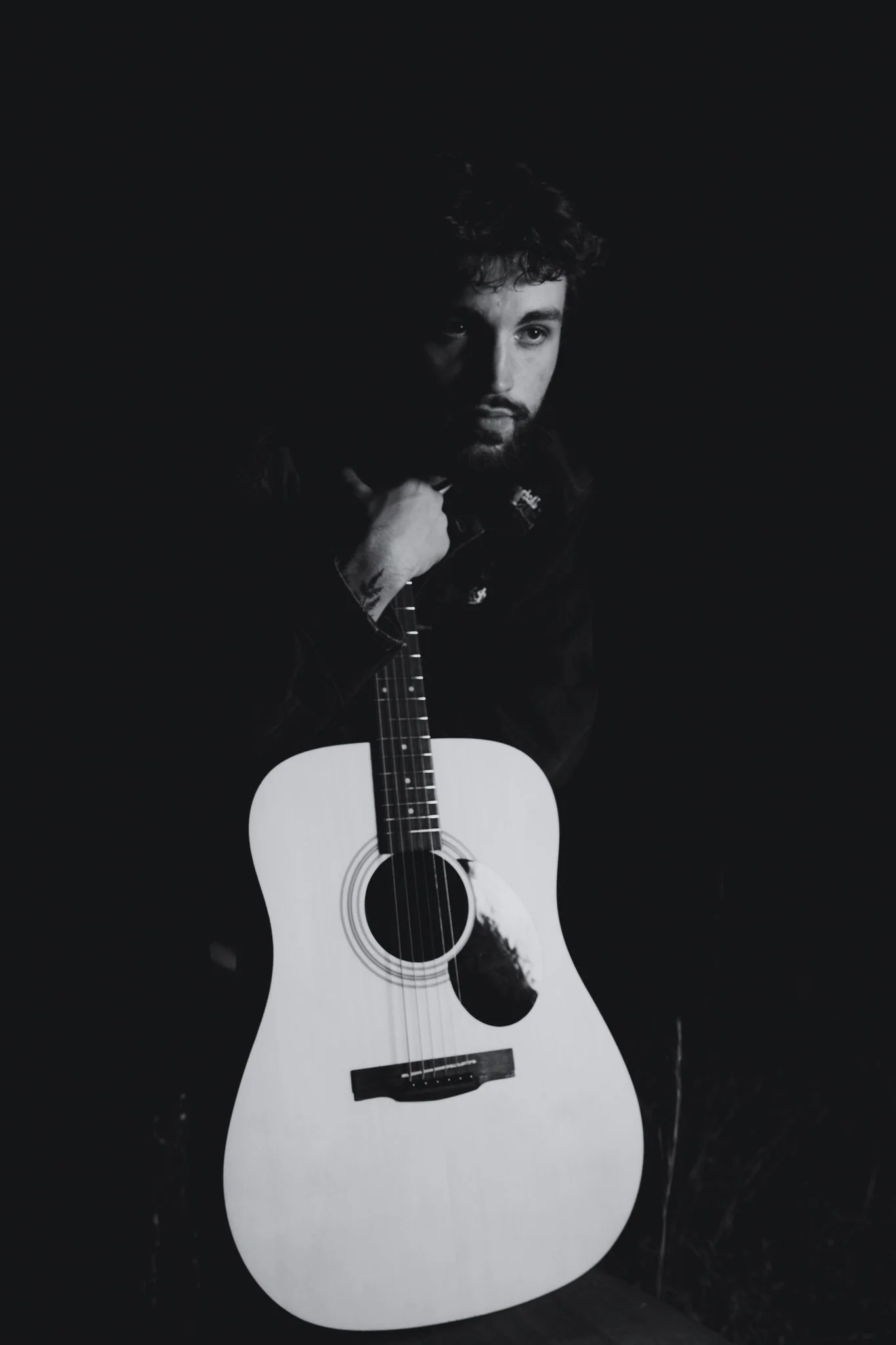 Black and white photo of a man with dark curly hair and a beard, holding an acoustic guitar with a light-colored body, against a plain black background.