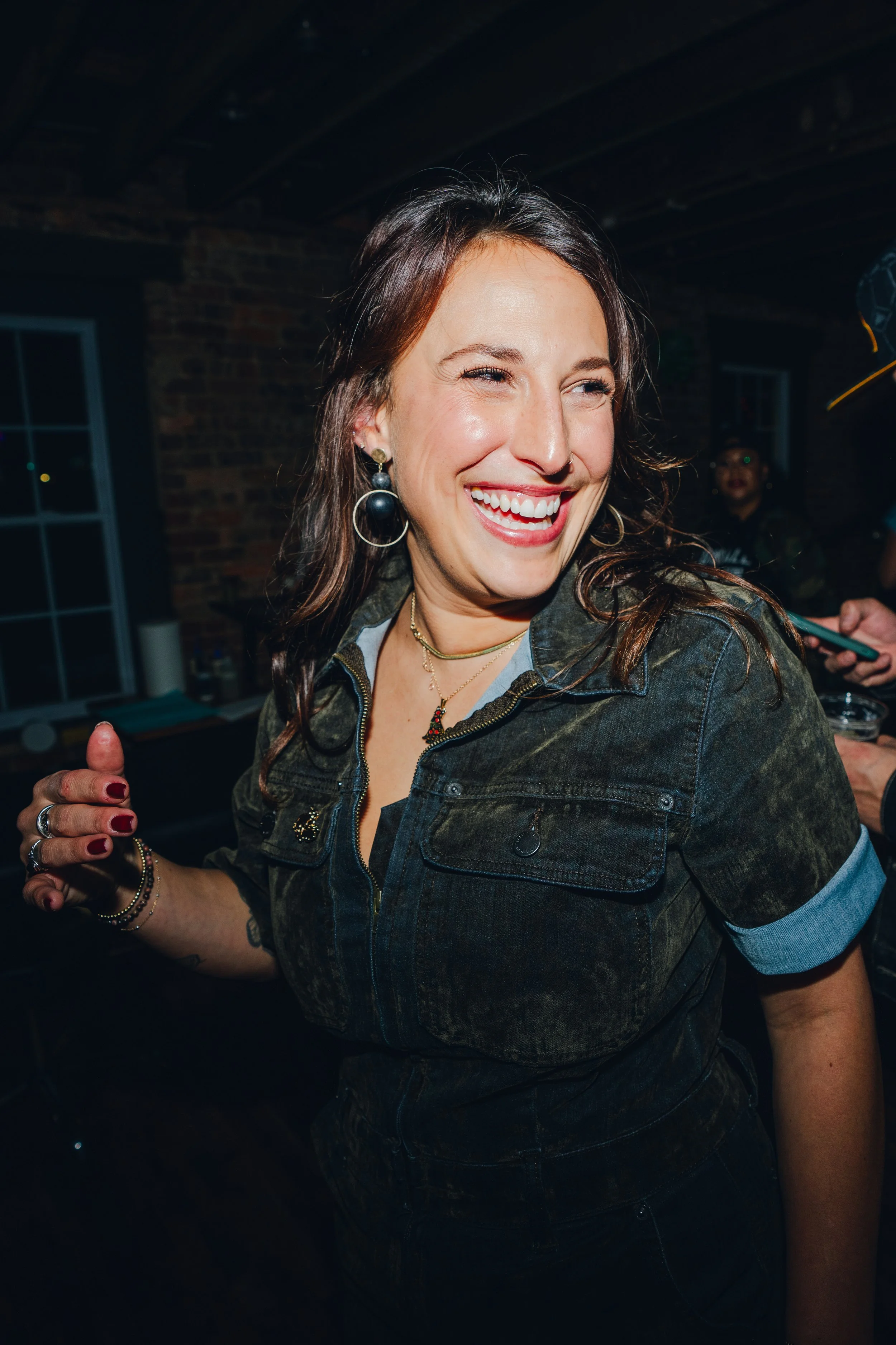 A woman smiling and laughing, wearing a dark denim jacket, earrings, rings, and layered necklaces, in a lively indoor setting with brick walls and windows.