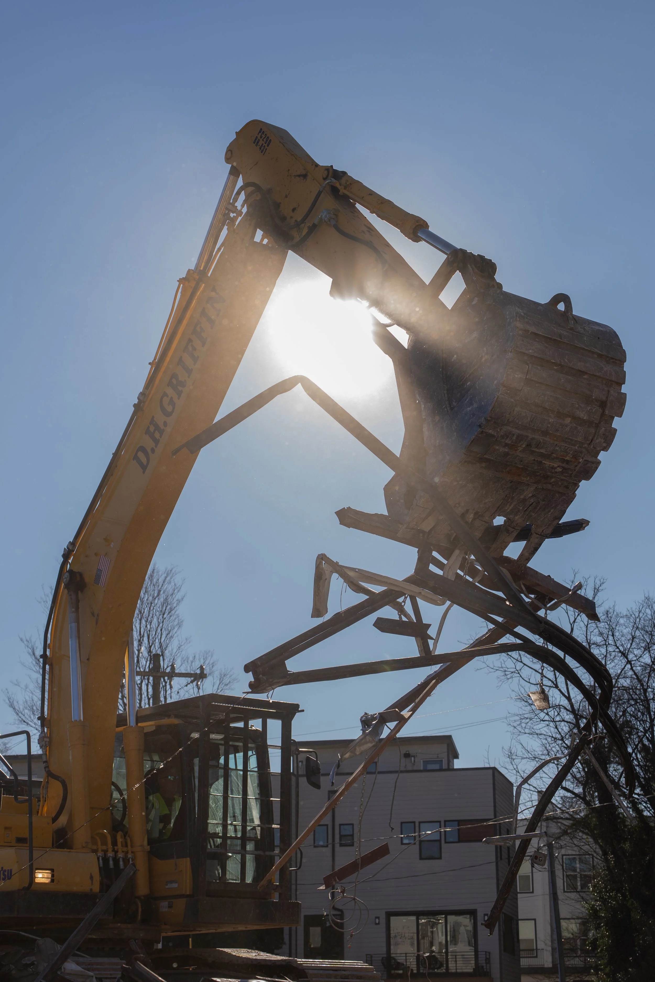 A construction excavator with a bucket is lifting debris and broken wood in a residential area with houses in the background against a clear blue sky.