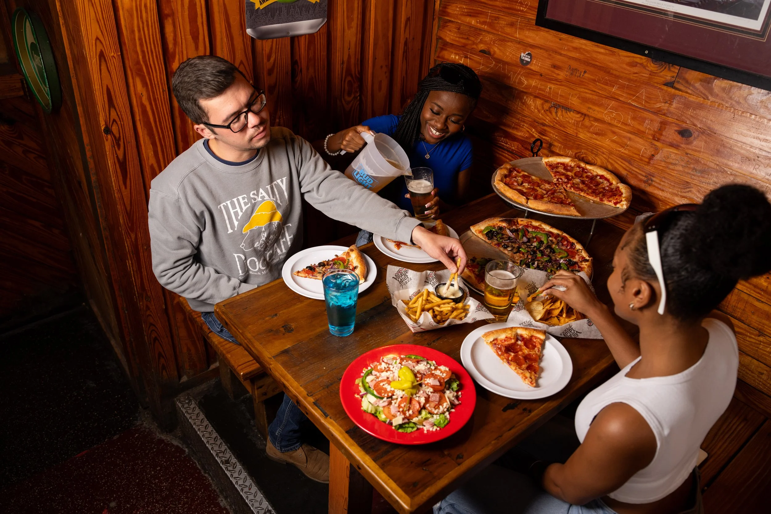 Four people sitting at a wooden table inside a restaurant, enjoying pizza, with drinks and a salad.