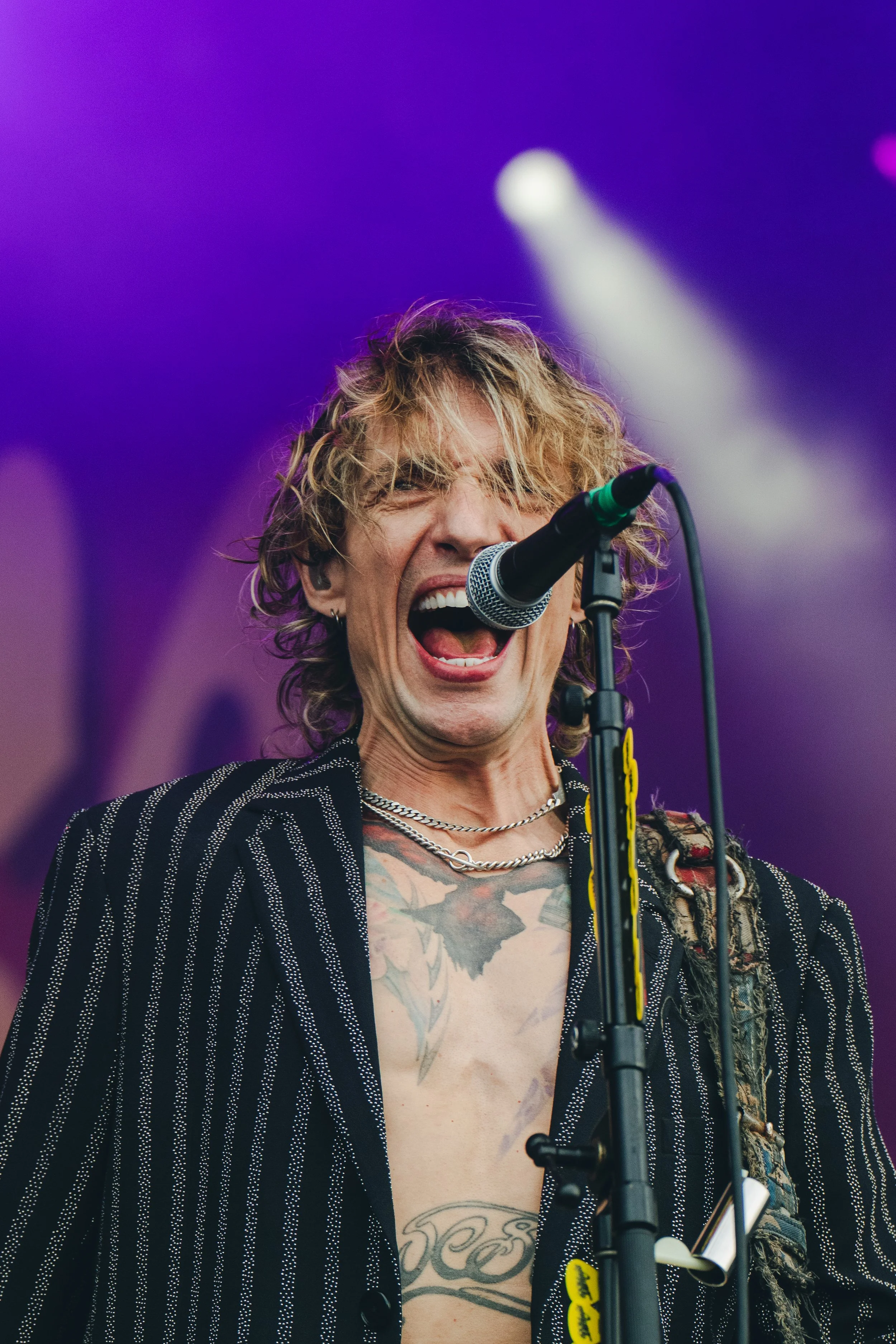 A young male singer with curly blond hair passionately performing on stage, singing into a microphone with purple stage lights in the background.