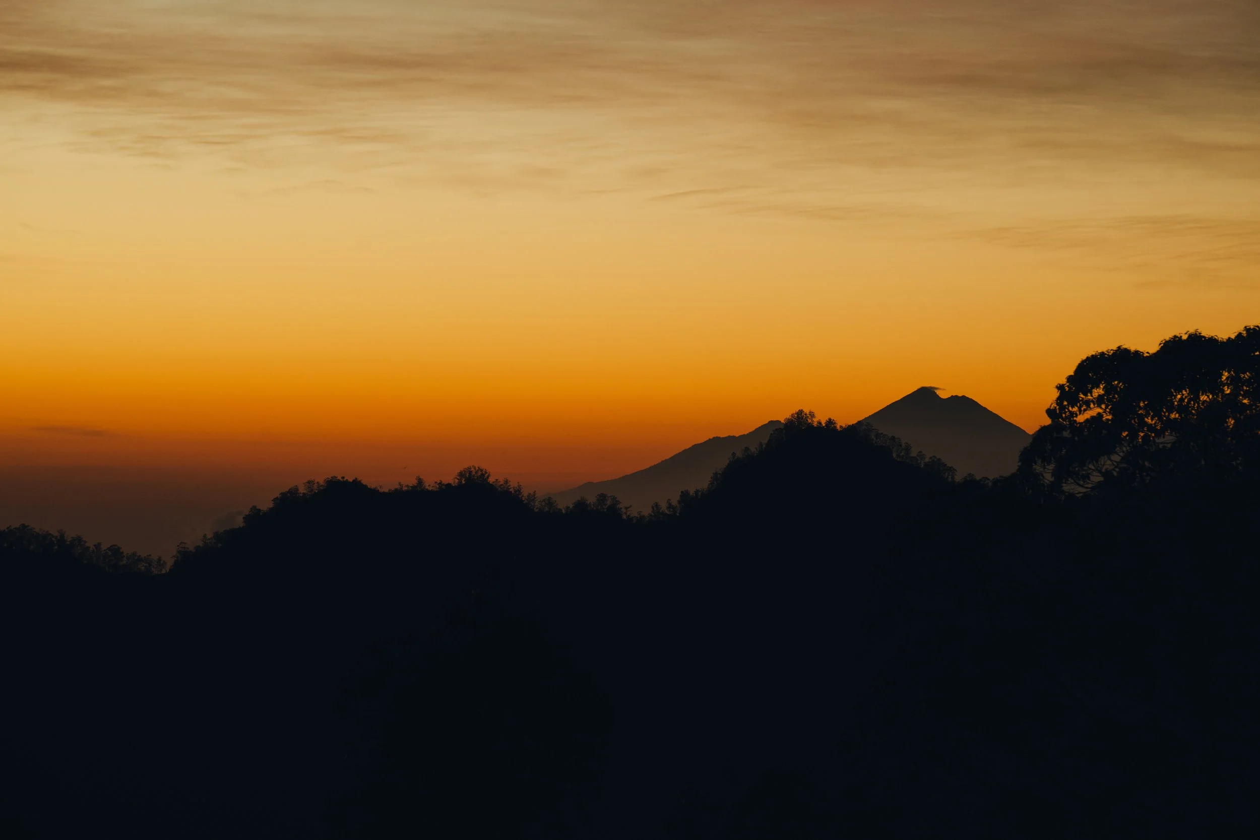 A silhouette of a mountain and trees against a sunset sky with orange and yellow hues.