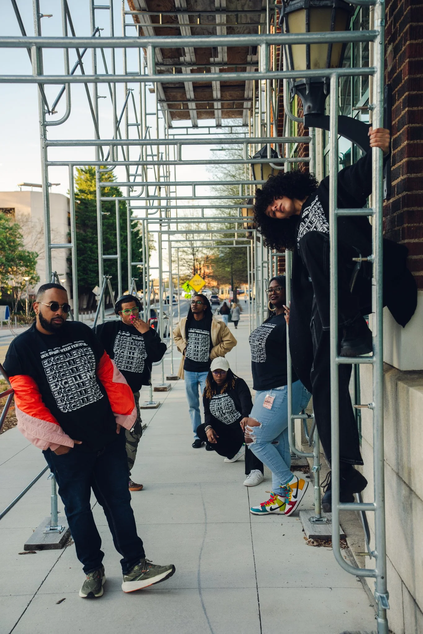 A group of people wearing matching black shirts with white text, standing on a sidewalk next to a metal scaffold and a brick wall, with some trees and a street in the background.