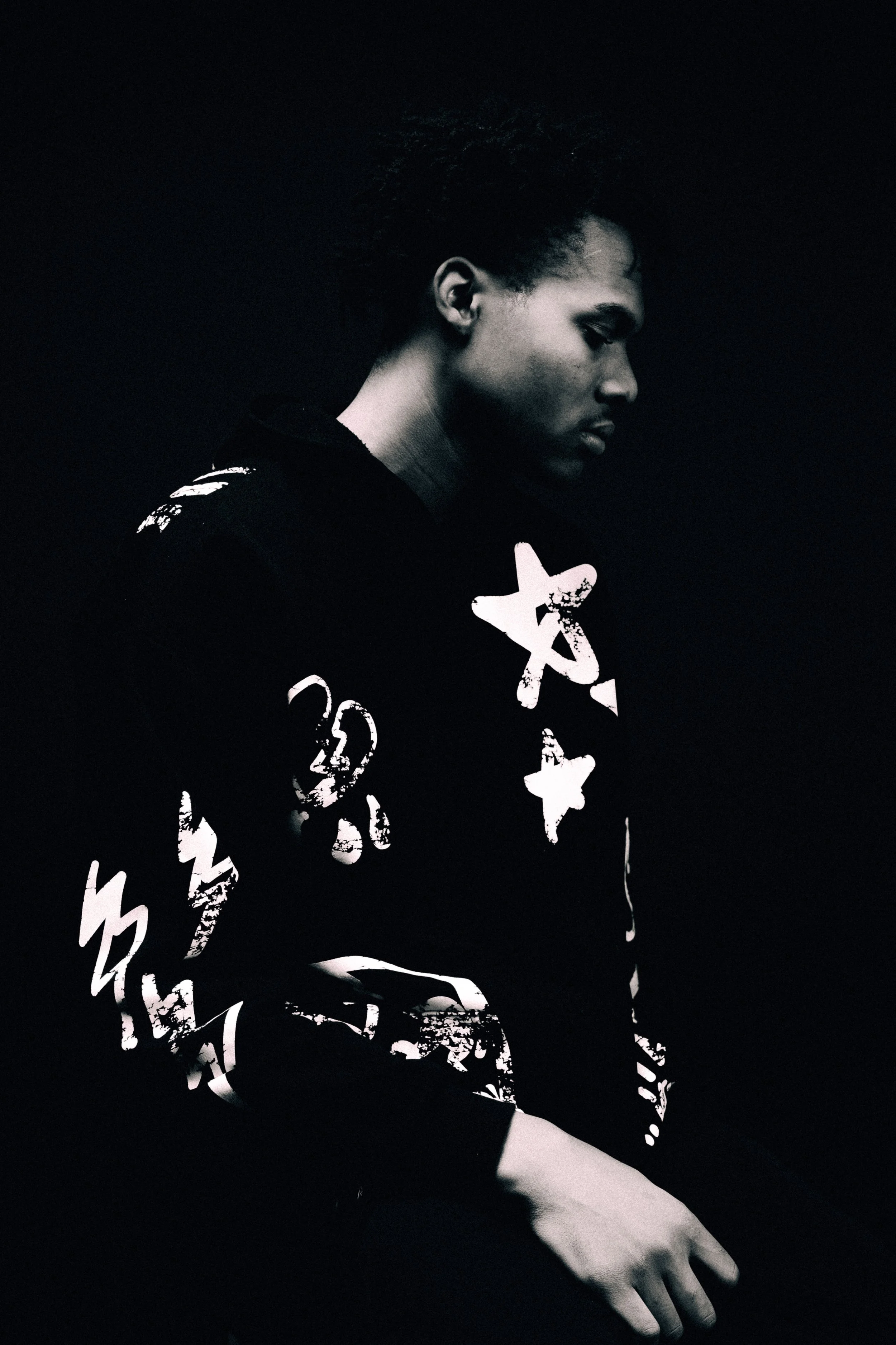 Black and white portrait of a young man with short curly hair, looking down, wearing a dark sweatshirt with graphic white text.