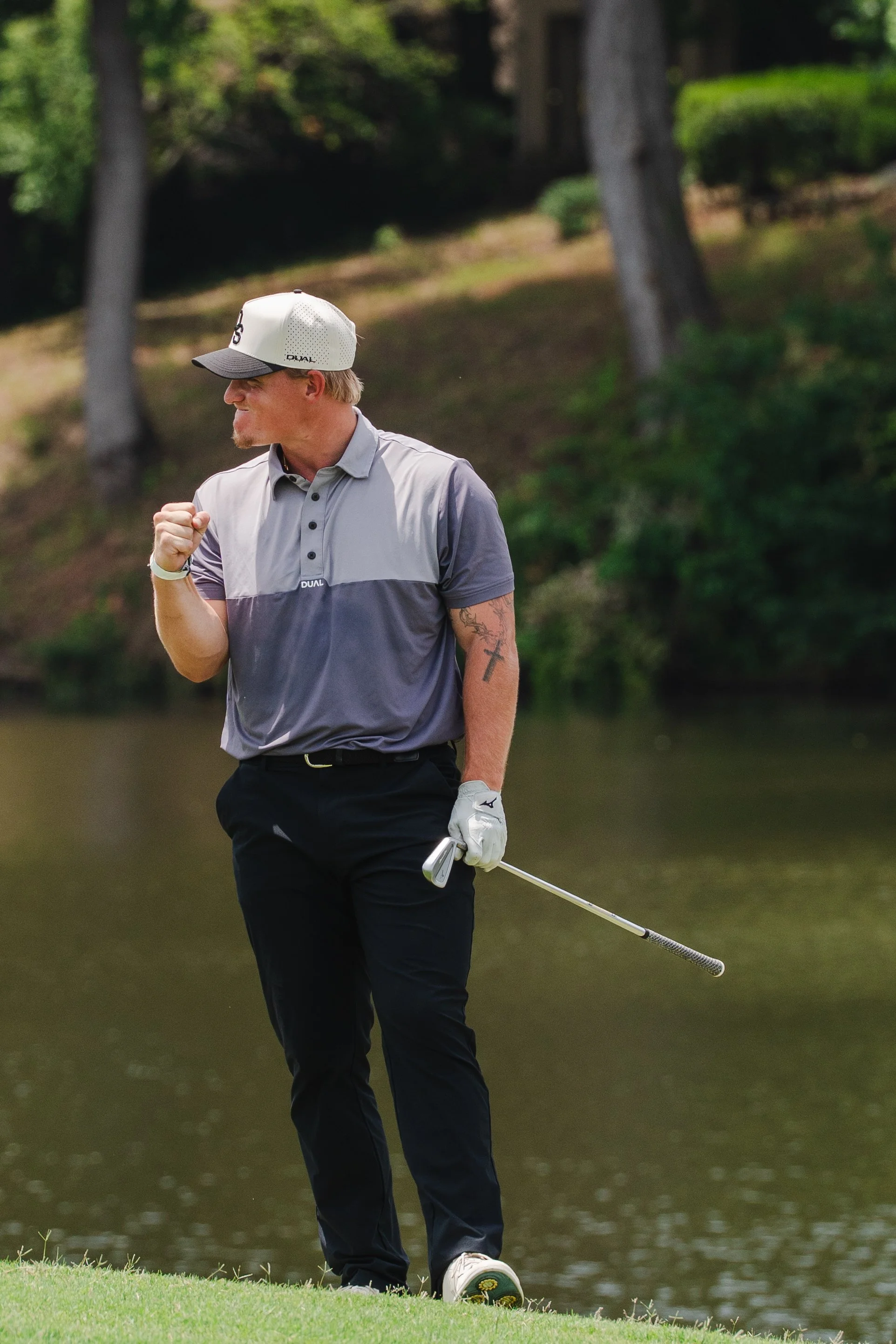 A man in a golf outfit celebrating on a golf course near a water hazard, with trees in the background.