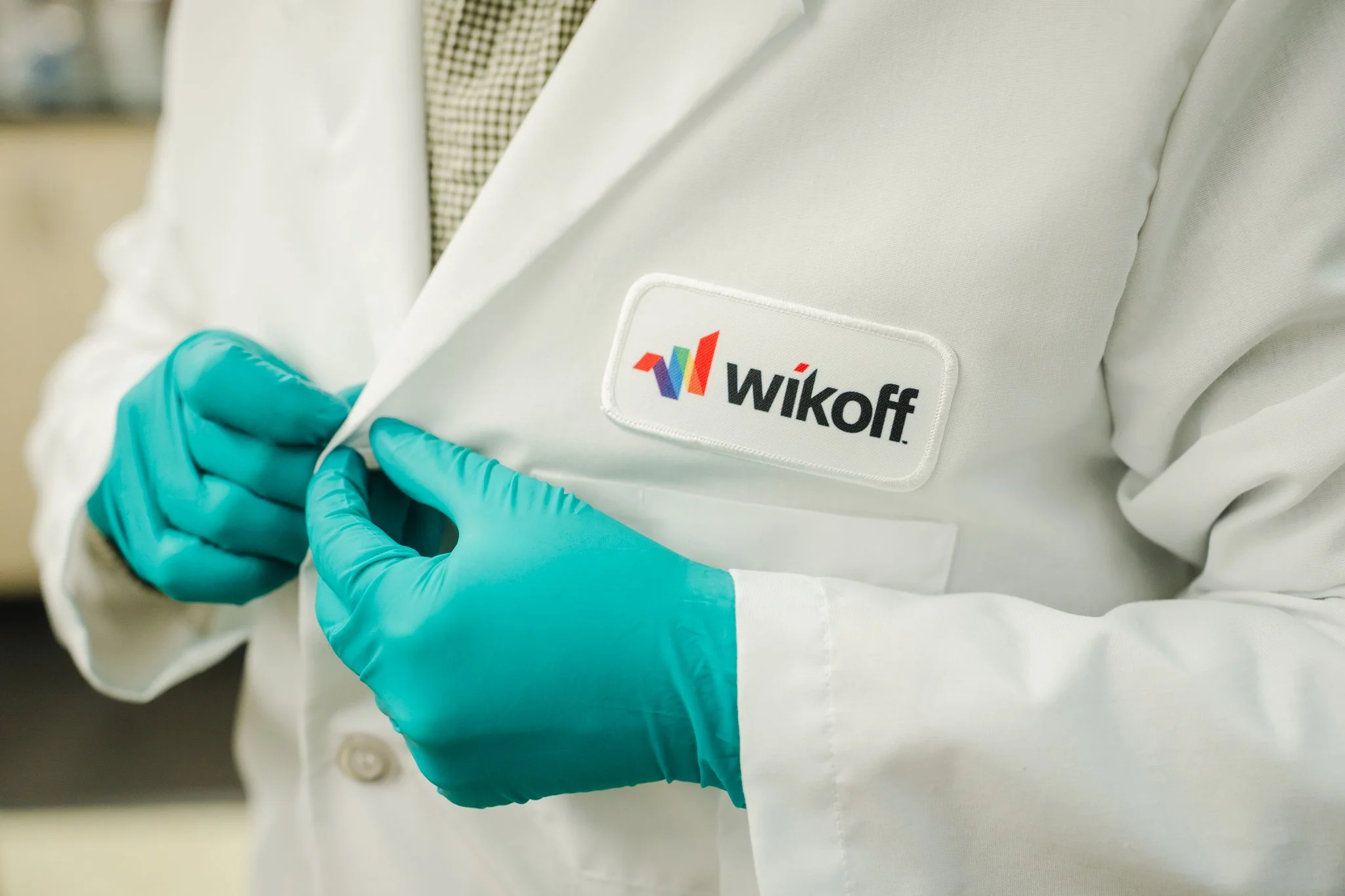 Close-up of a person wearing a white lab coat with a WiKoff logo and sporting teal gloves, with hands crossed.
