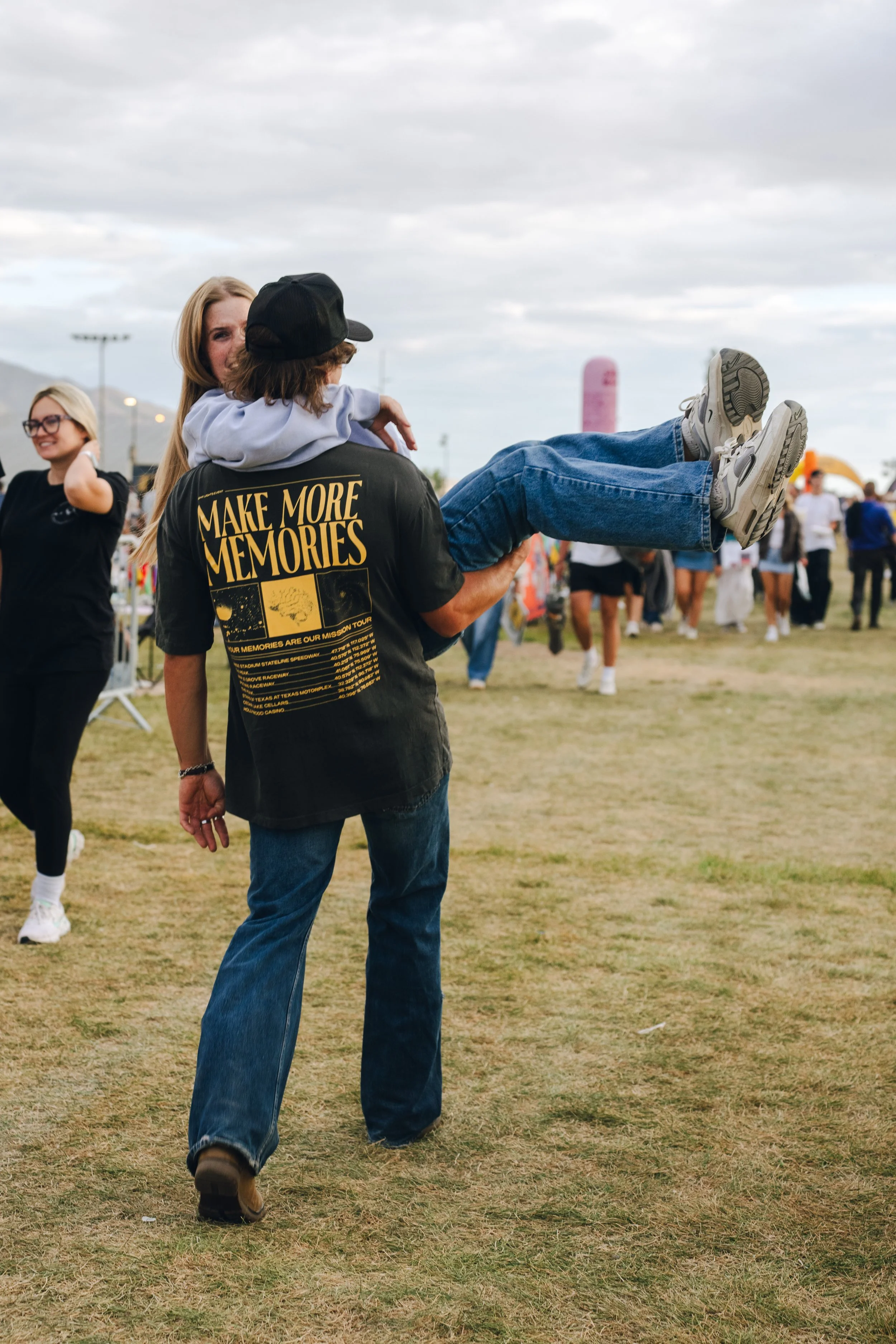 A person is carrying a child in their arms at an outdoor event or fairground, with other people and tents in the background. The person is wearing a black shirt with yellow text that says "Make More Memories" and has long hair. The child is wearing a