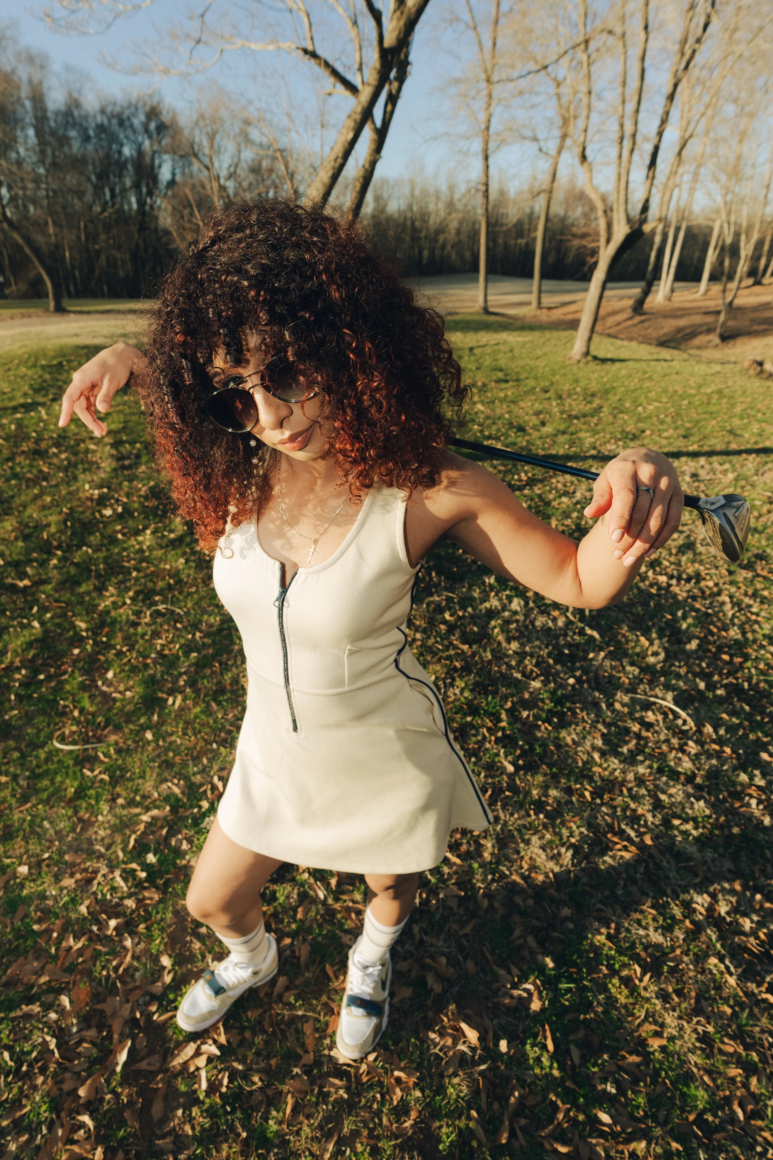 A woman with curly hair and sunglasses in a cream-colored dress outdoors on a sunny day, holding a golf club over her shoulders, standing on a grassy area with trees in the background.