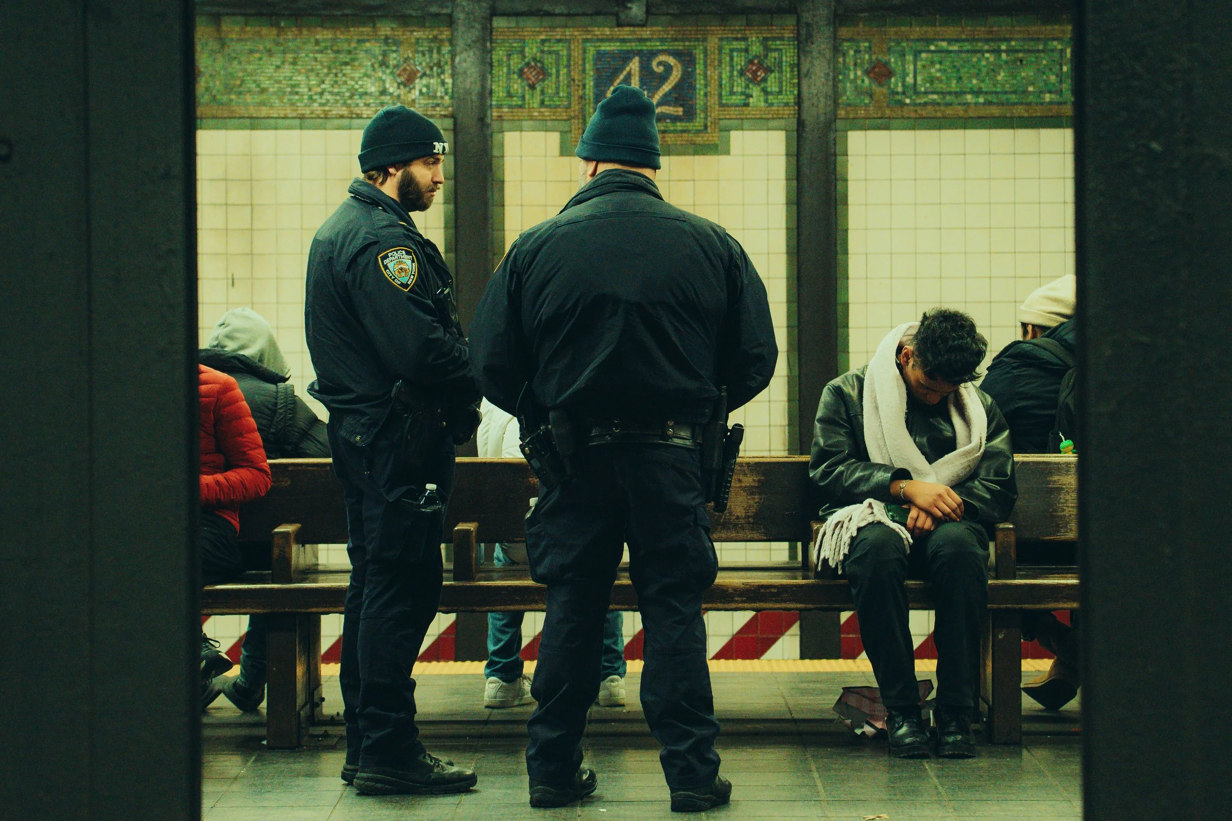 Two police officers in black uniforms and beanies standing and talking in a subway station. Behind them, people are sitting on benches, some looking down or at their phones. The station has tiled walls and a sign with the number 42.